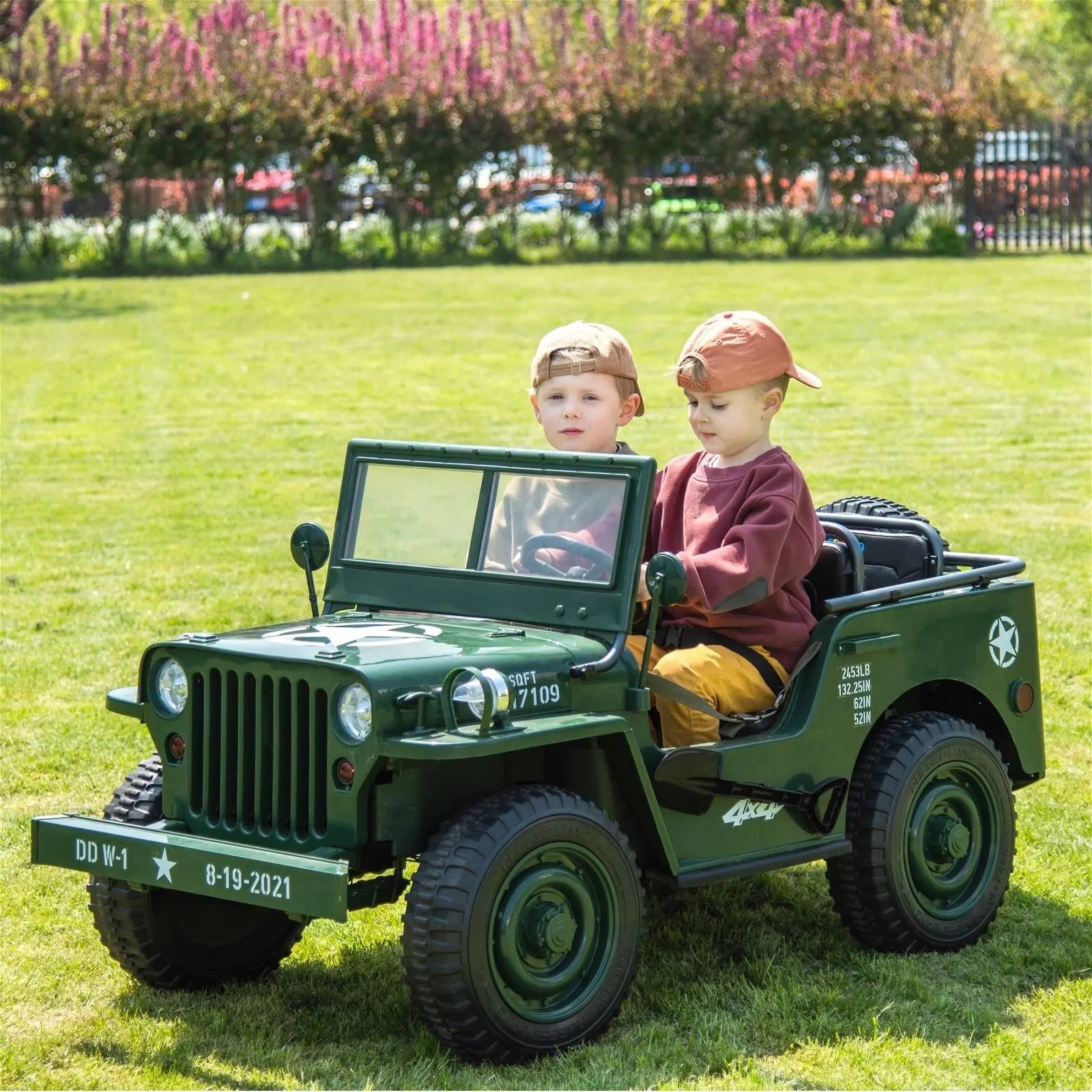 Two kids riding a green 3-seater electric military jeep toy outdoors on grass