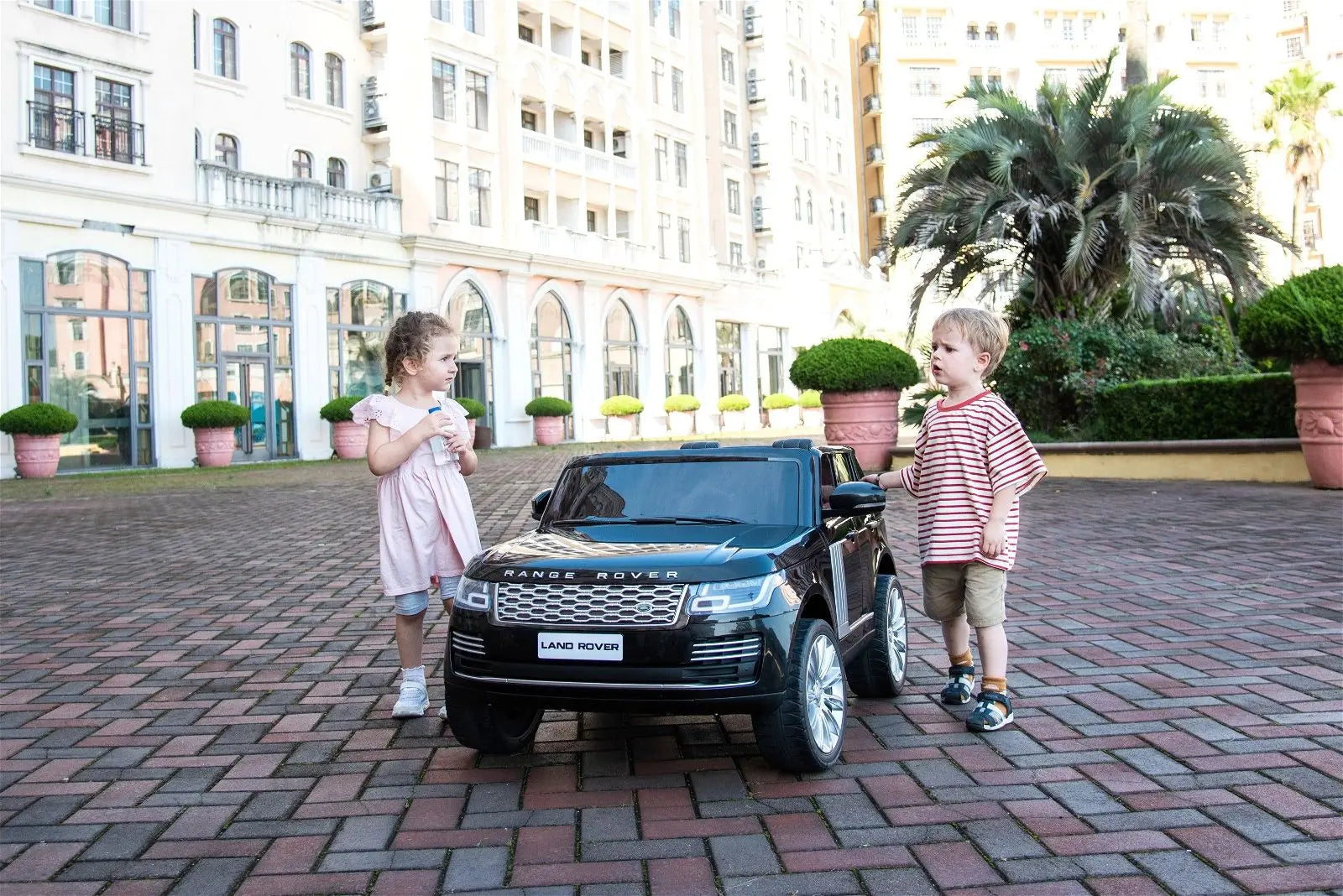Kids standing by a black 2-seater Range Rover ride-on car outdoors on brick pavement
