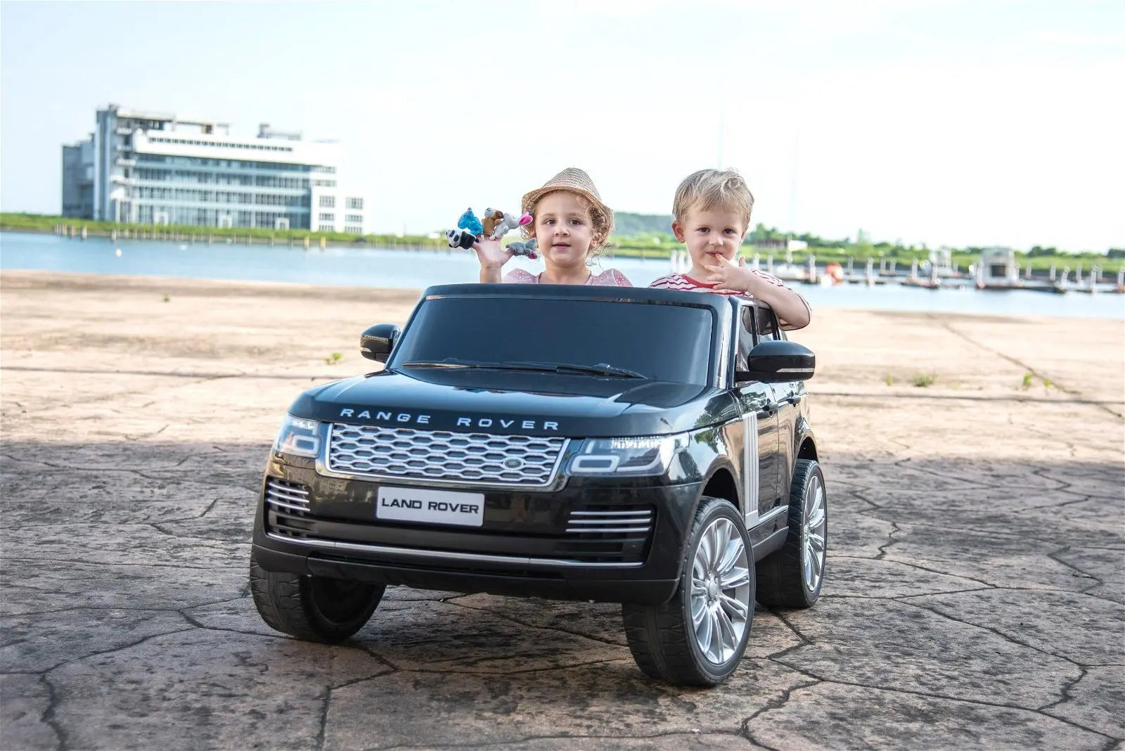 Two kids riding in a black 2-seater Range Rover ride-on toy car outdoors by water