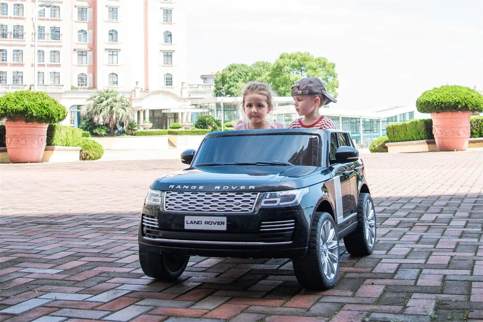 Two kids in a black Range Rover 2-seater ride-on toy car outdoors on brick pavement