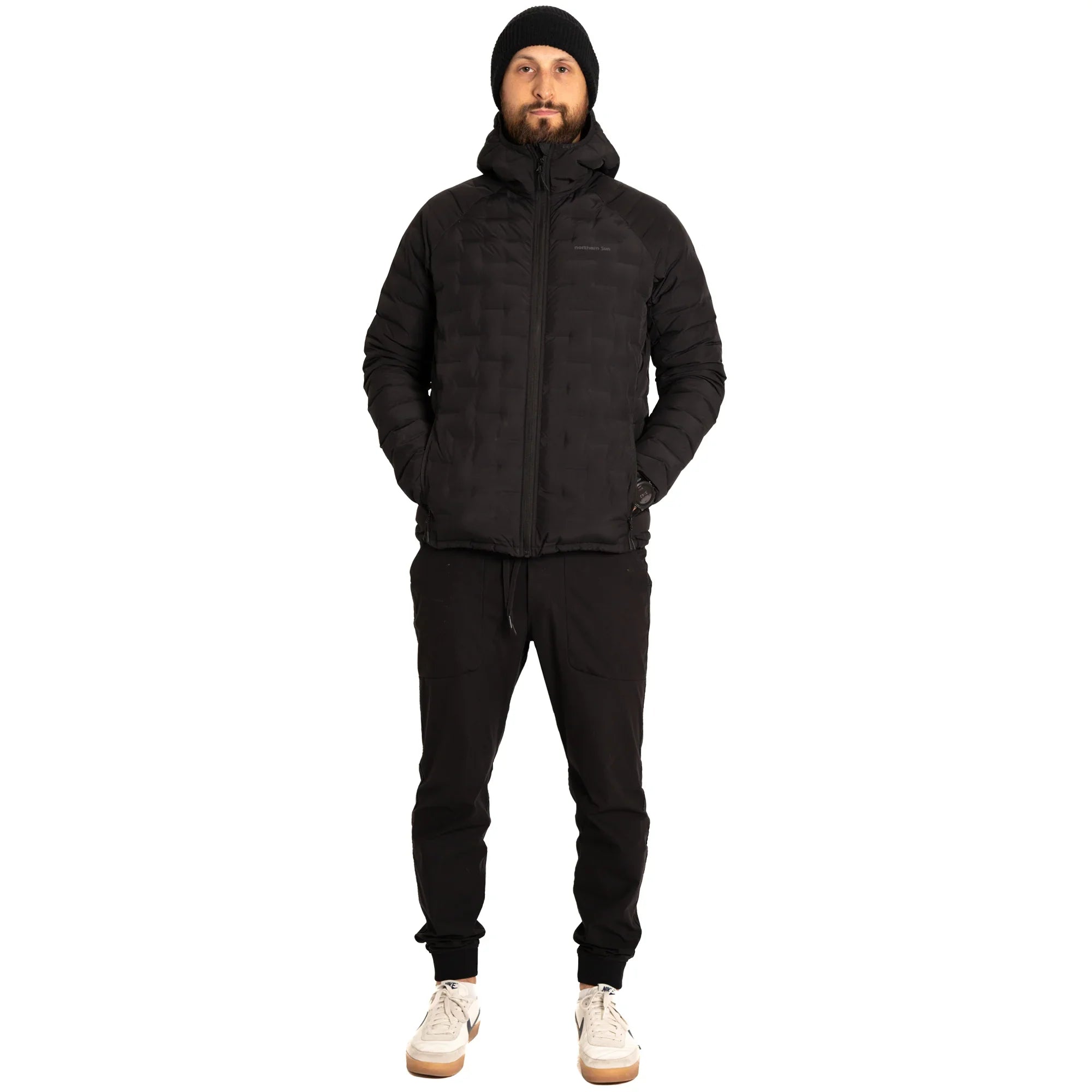 Man wearing black puffer jacket, black joggers, beanie, white sneakers on white background