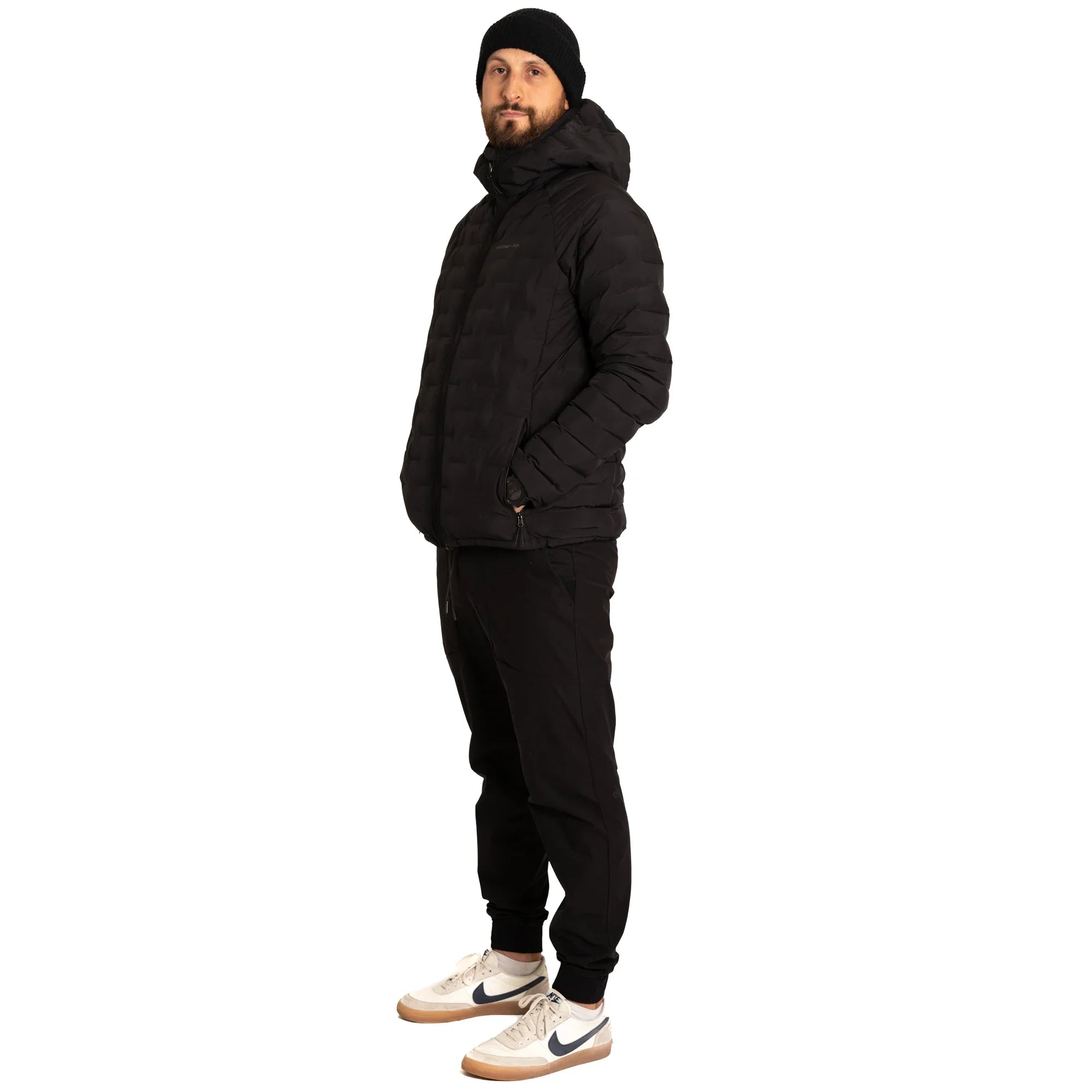 Man wearing black puffer jacket, black joggers, beanie, and Nike sneakers on white background