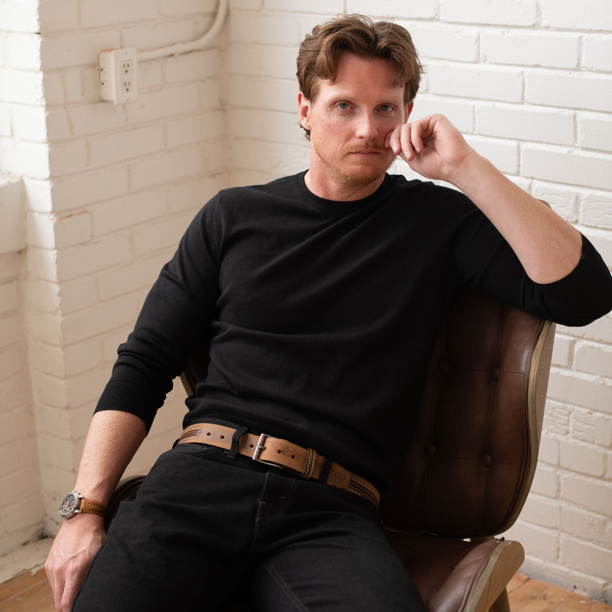 Man in black long sleeve shirt and wristwatch sitting on brown chair in modern indoor setting