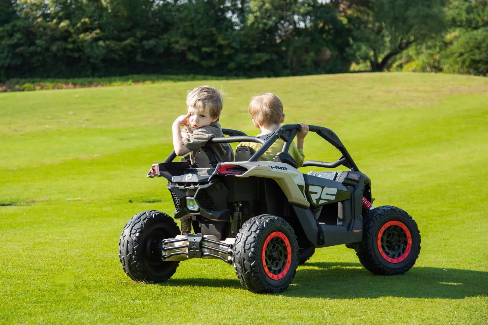 Two children riding a 2-seater electric Can-Am Maverick UTV toy on grass outdoors.