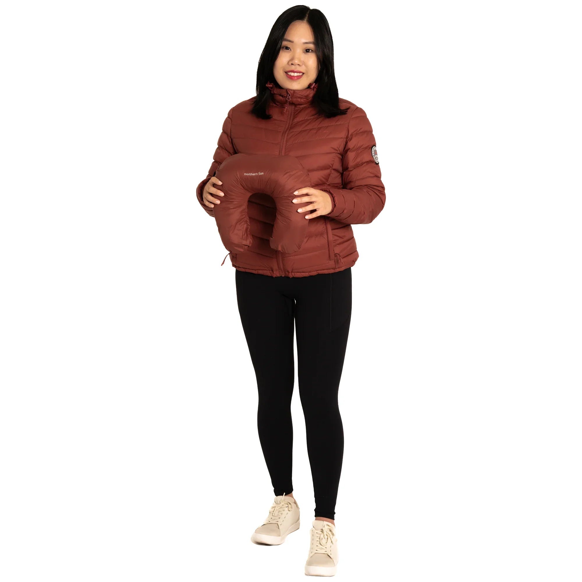 Woman in brown puffer jacket holding brown travel neck pillow, black leggings, white shoes