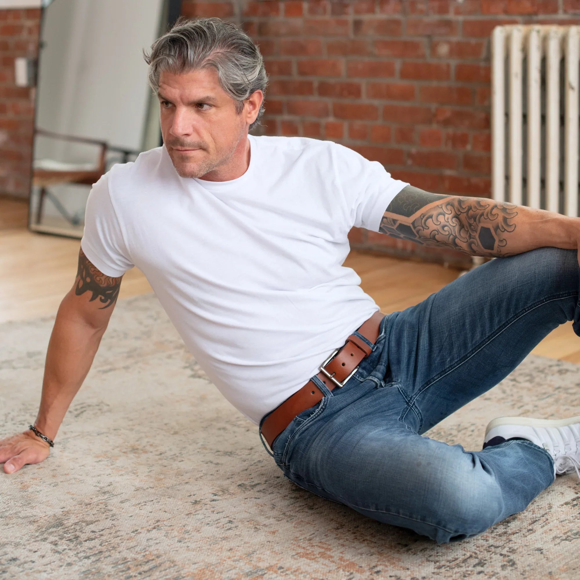 Man in white t-shirt and jeans with arm tattoos sitting on rug in modern indoor setting