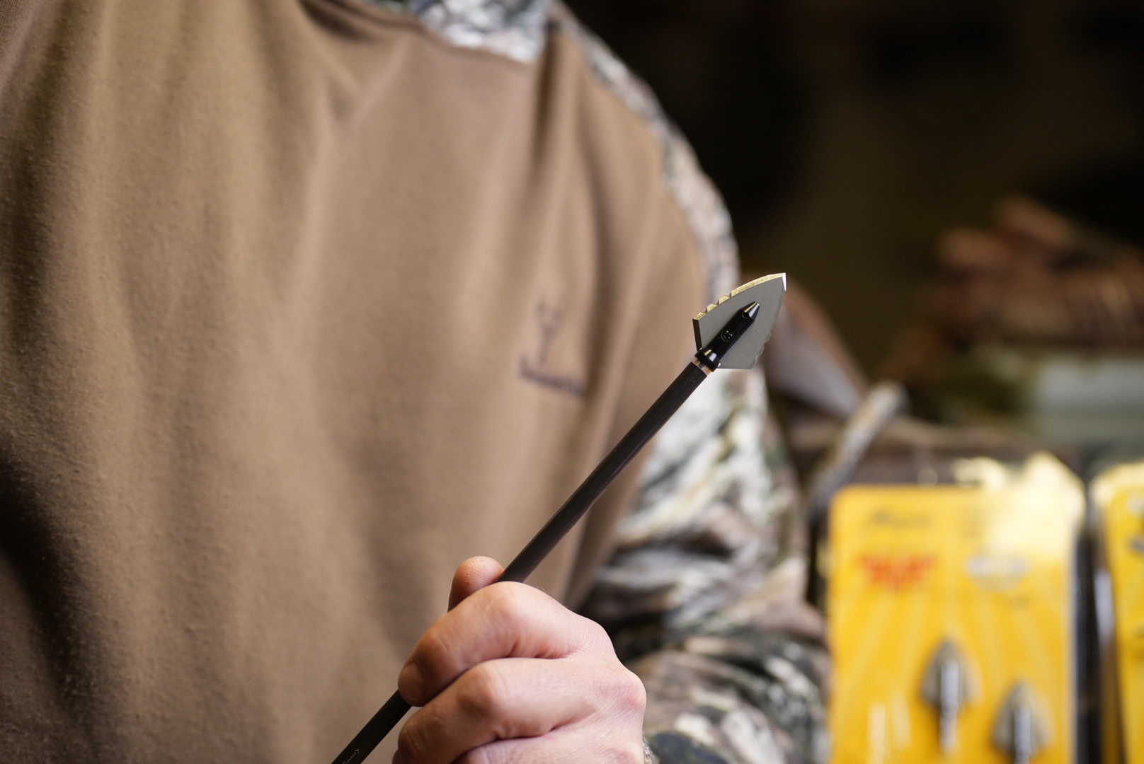 Hand holding hunting arrow with broadhead tip, archery gear in shop background