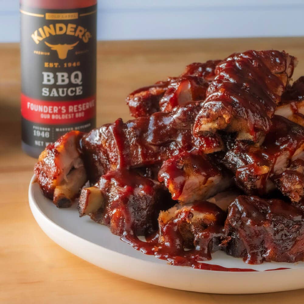 Plate of BBQ pork ribs covered in barbecue sauce with Kinder's BBQ sauce bottle in background