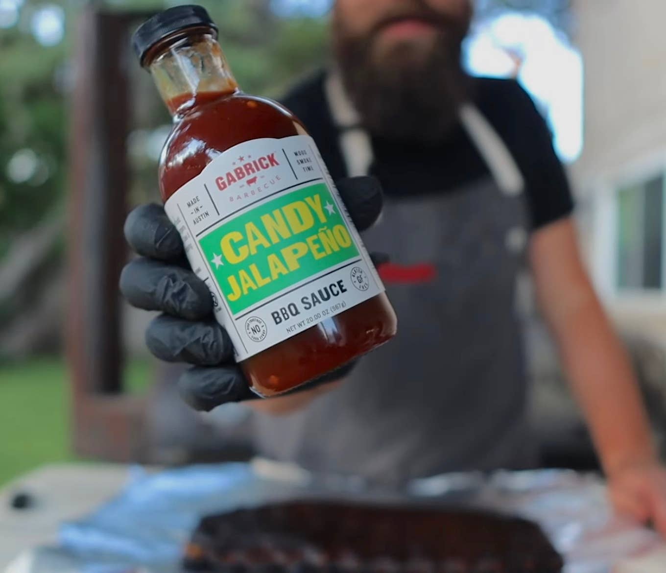 Person wearing gloves holding Gabrick BBQ Candy Jalapeño sauce bottle outdoors near grill