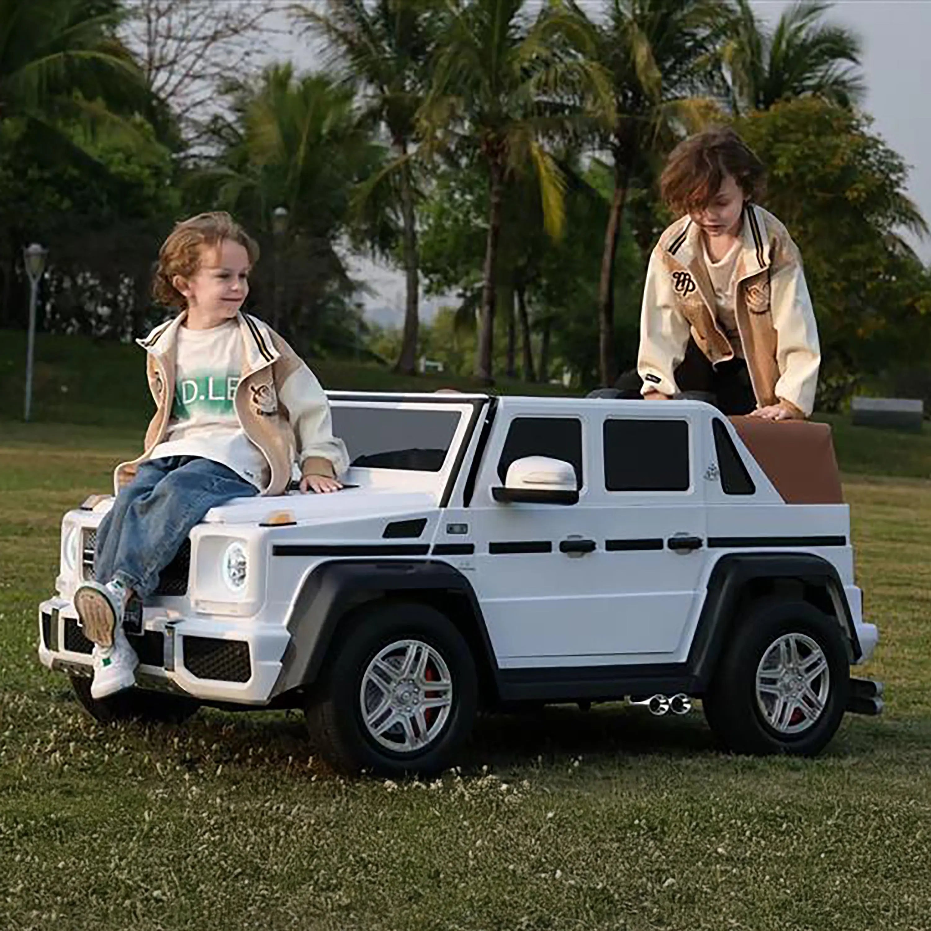 Children playing outdoors on a white ride-on toy SUV in a grassy park with palm trees.