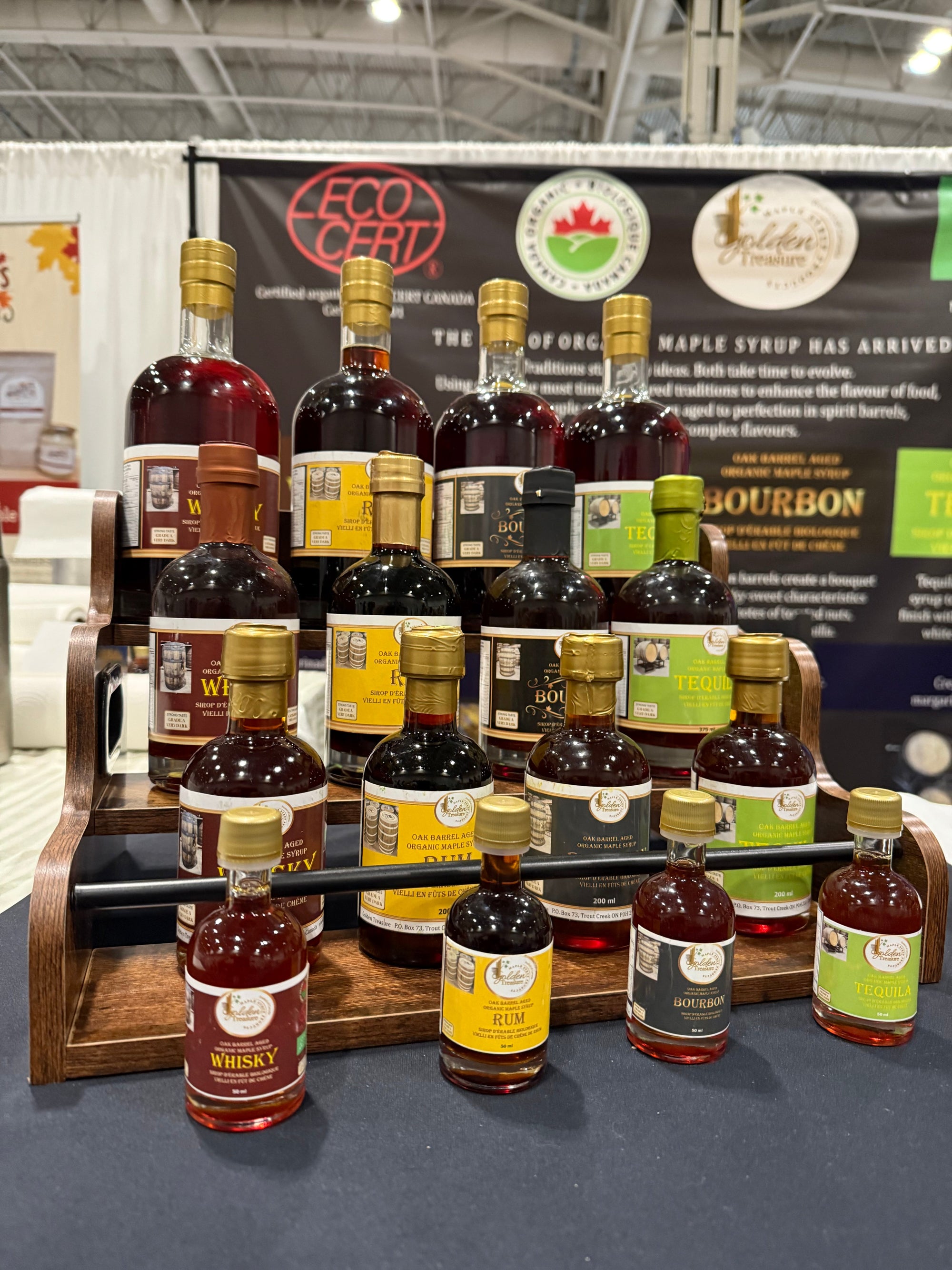 Display of assorted oak barrel aged maple syrup bottles with whisky, bourbon, rum, and tequila flavors at a trade show booth.