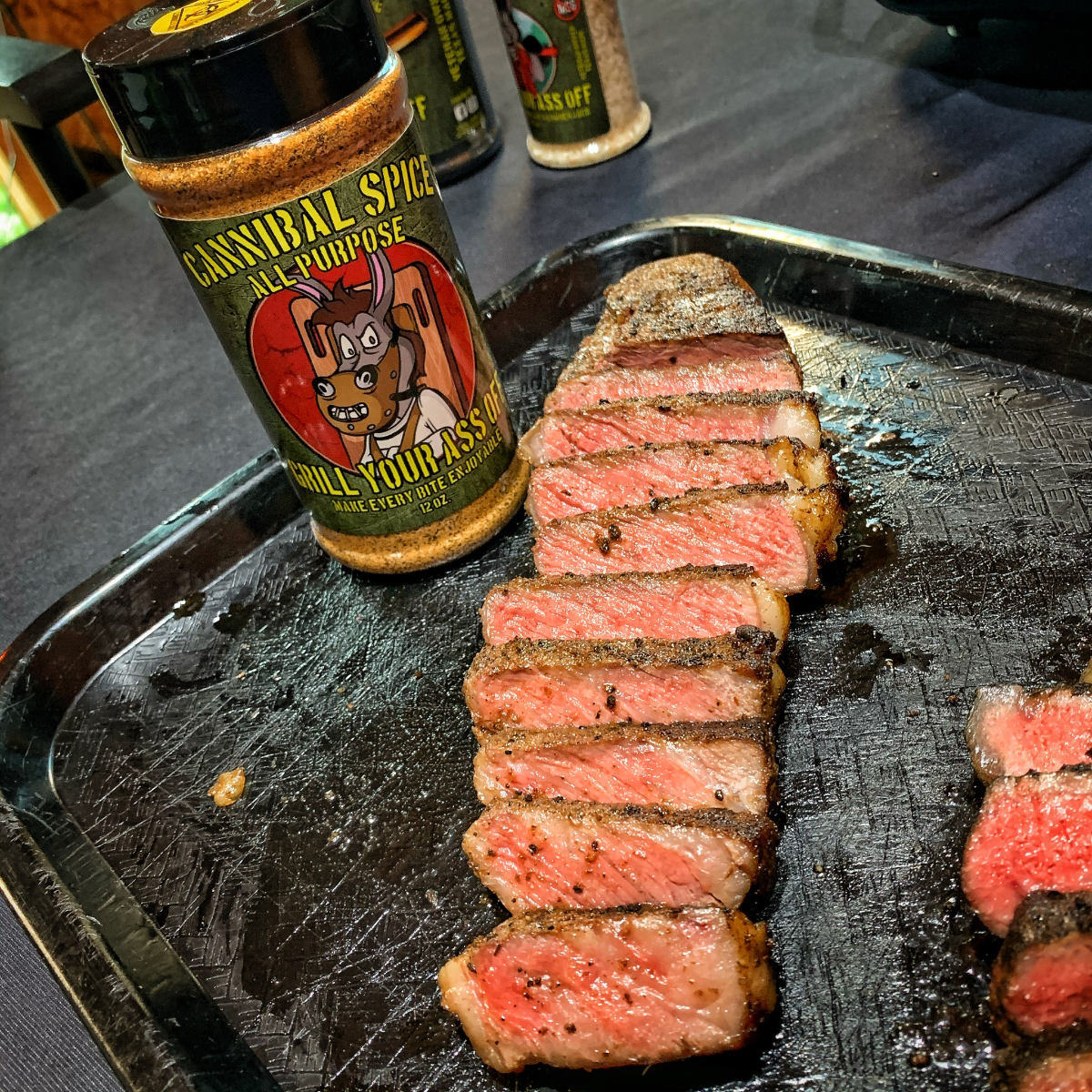 Grilled sliced steak on a black tray beside all-purpose seasoning bottle for outdoor cooking.