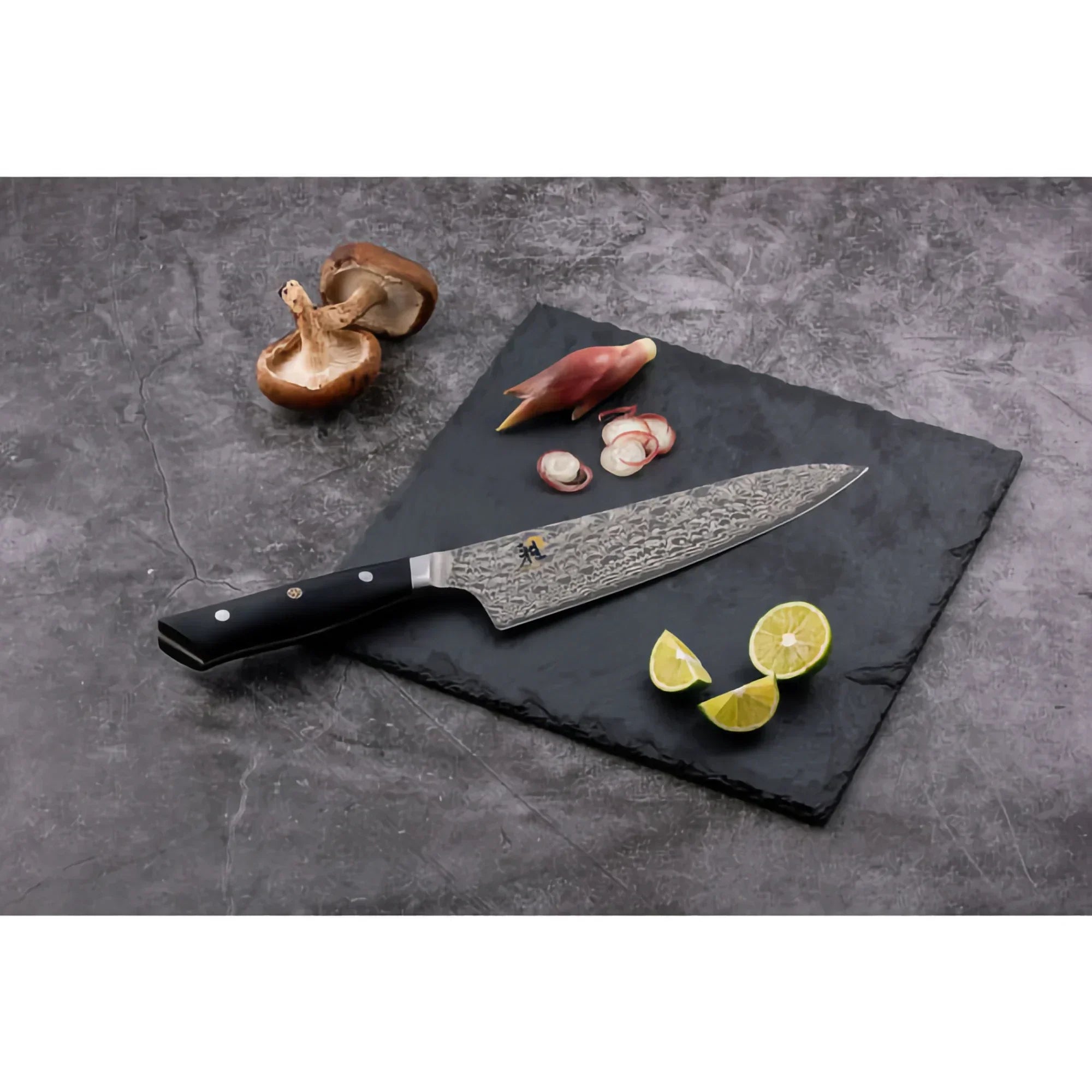 Damascus steel chef knife on slate board with mushrooms, sliced herbs, and lime wedges