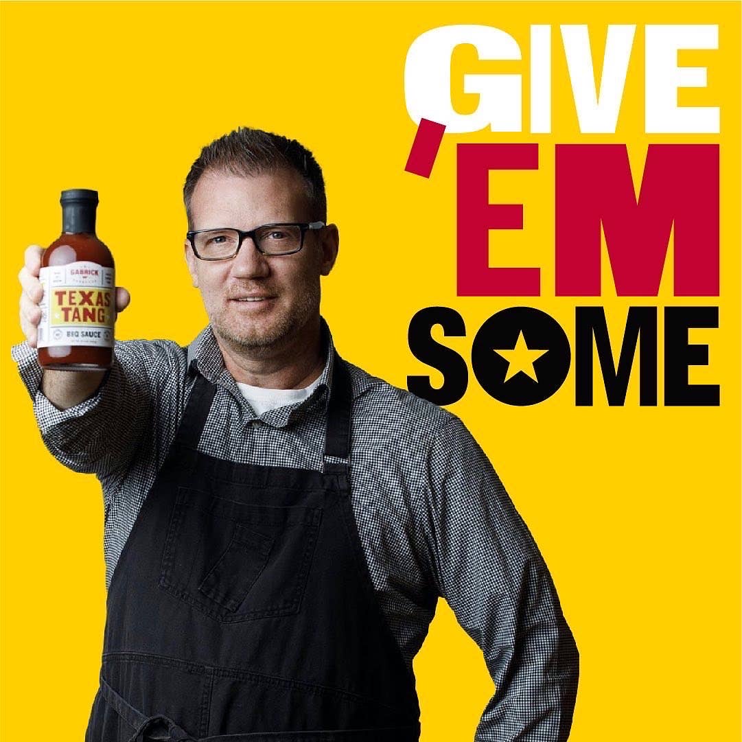 Man in apron holding Texas Tang BBQ sauce bottle on yellow background with 'GIVE 'EM SOME' text.