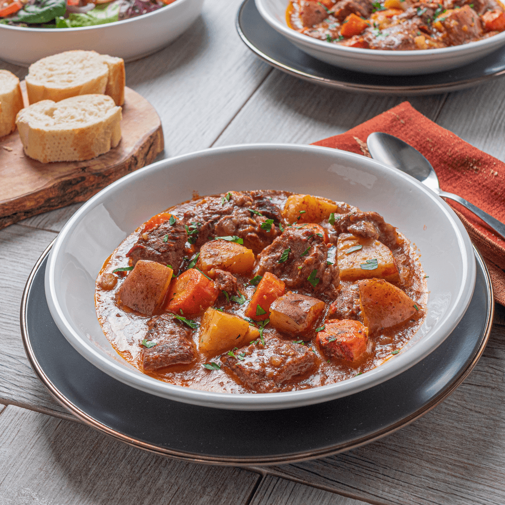 Hearty beef stew with potatoes and carrots in a bowl, served with bread and salad on table