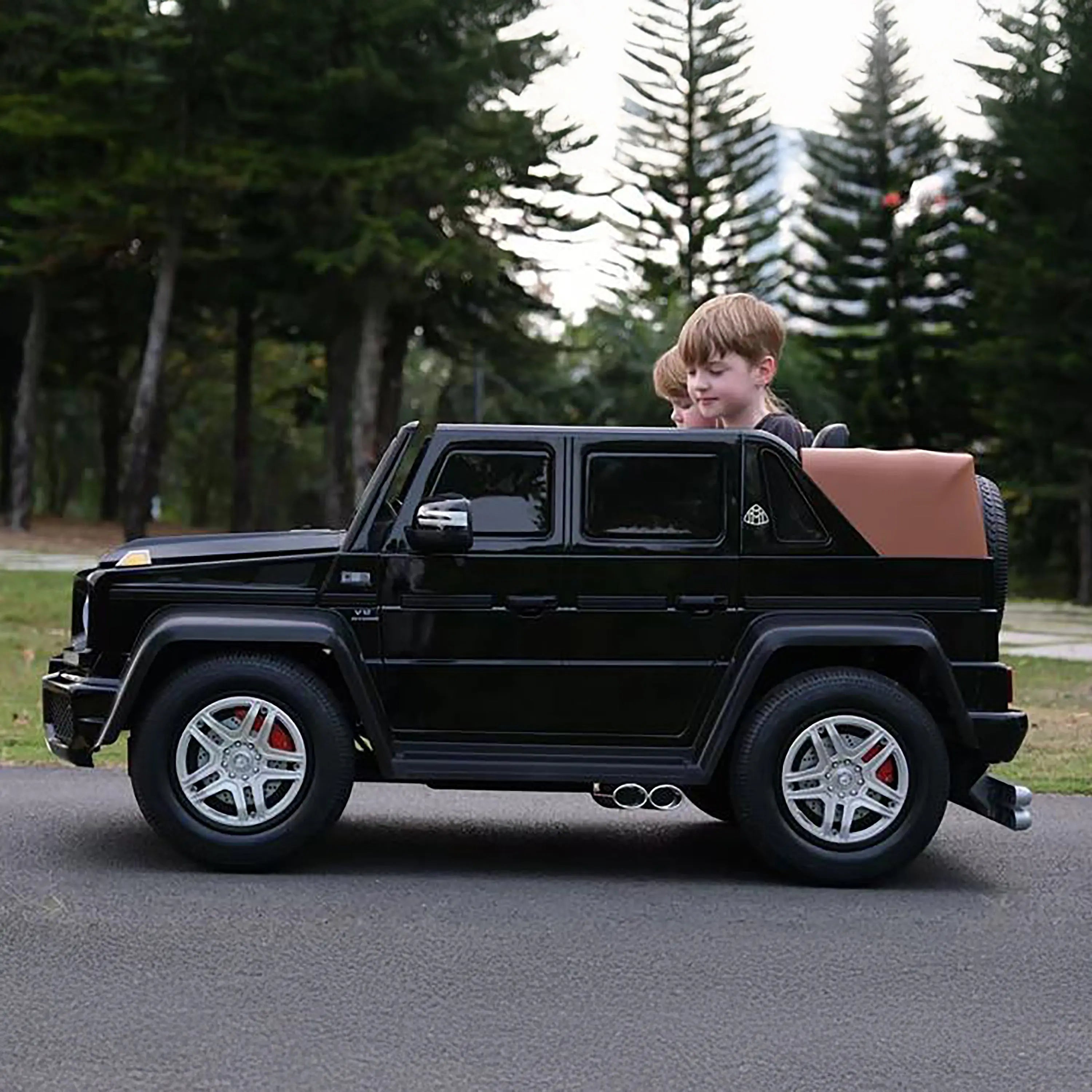 Two kids riding a black toy SUV outdoors on a paved road with trees in the background