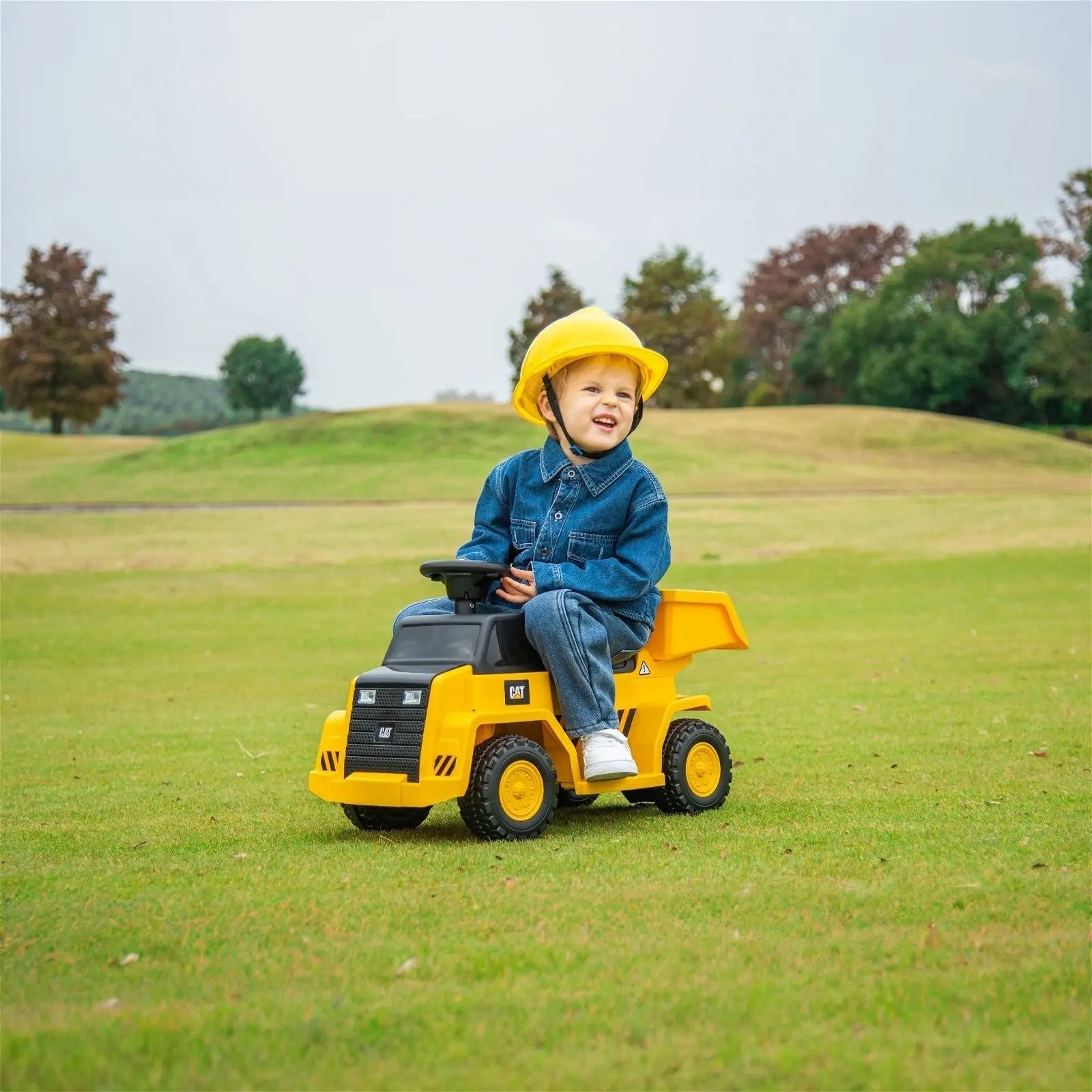 Child in denim jacket and yellow helmet rides yellow dump truck toy on grass outdoors
