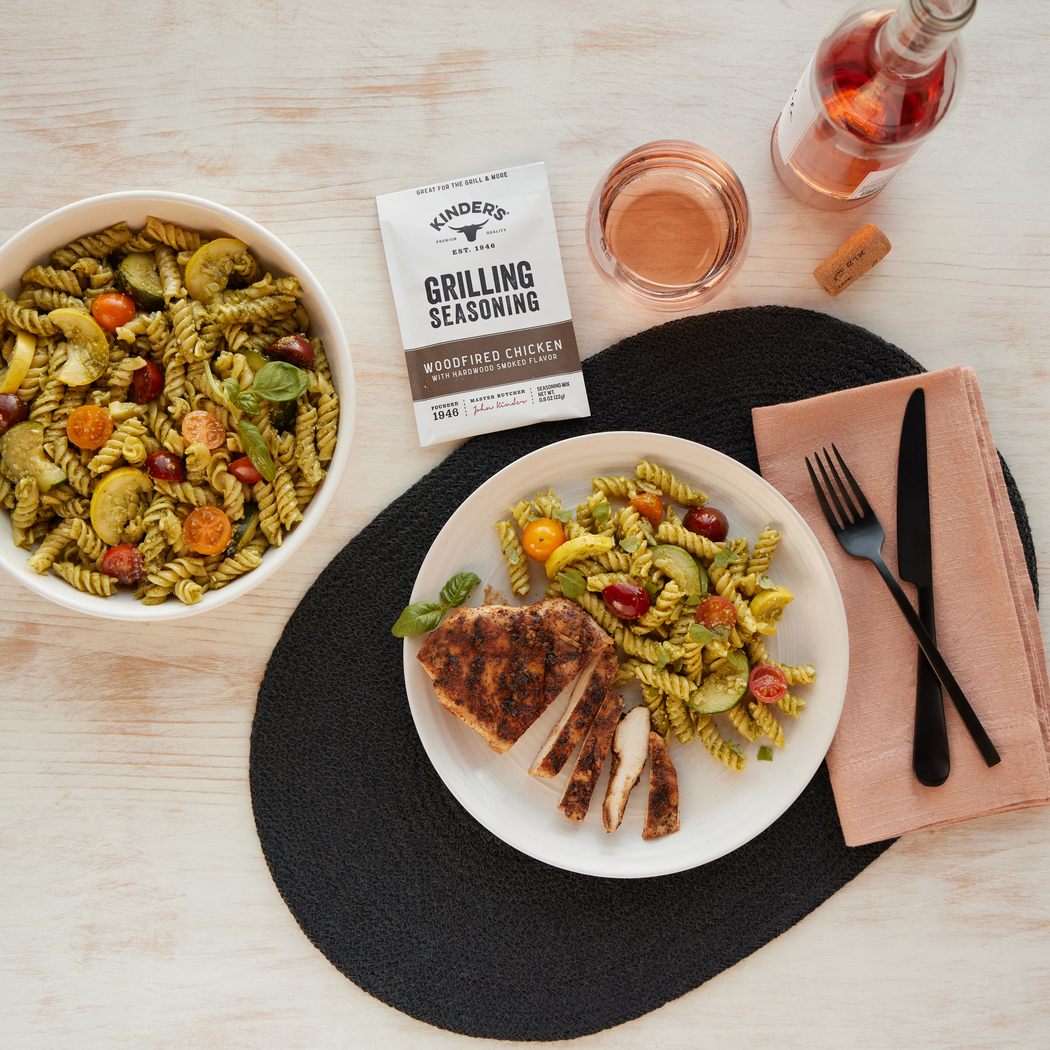 Pasta dish with grilled chicken on a plate, alongside a bowl of pasta and a bottle of rose wine on a wooden table.