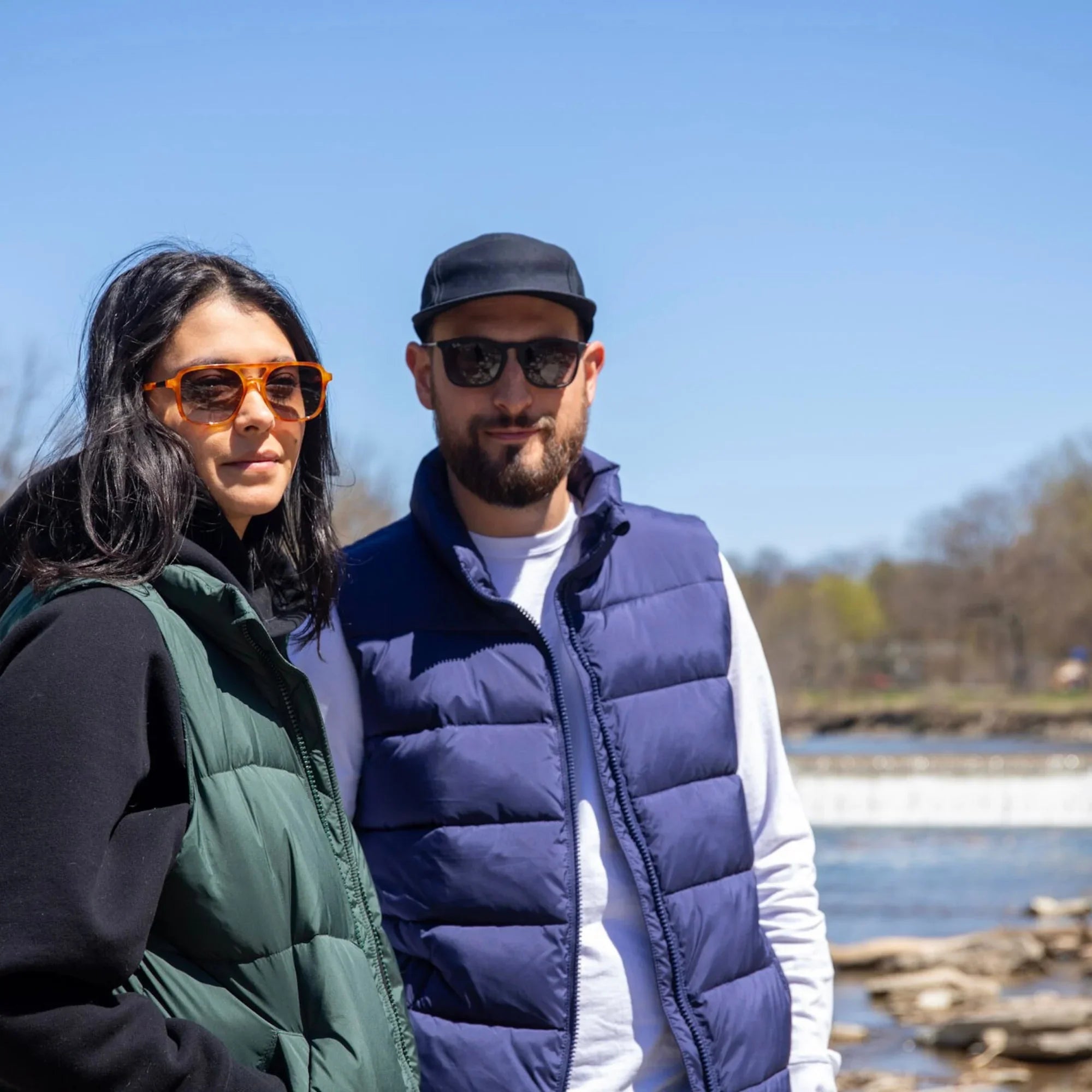 Man and woman outdoors in puffer vests and sunglasses by riverside on sunny day