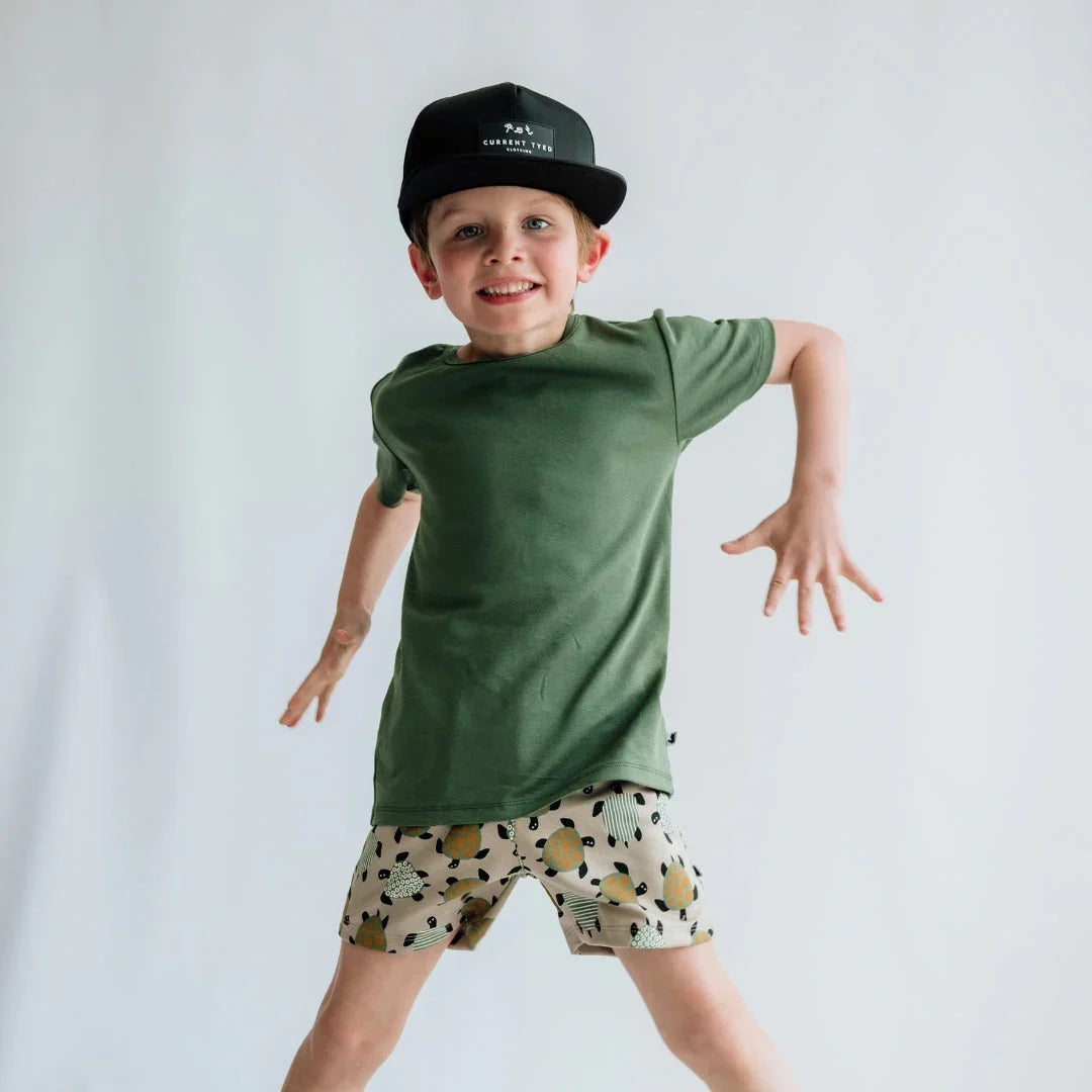 Smiling boy in olive green youth plain T-shirt and patterned shorts against white background