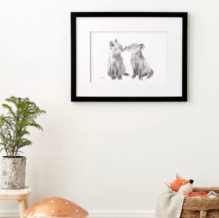 Framed sketch of two baby foxes on white wall, nursery decor with plant and plush fox toy