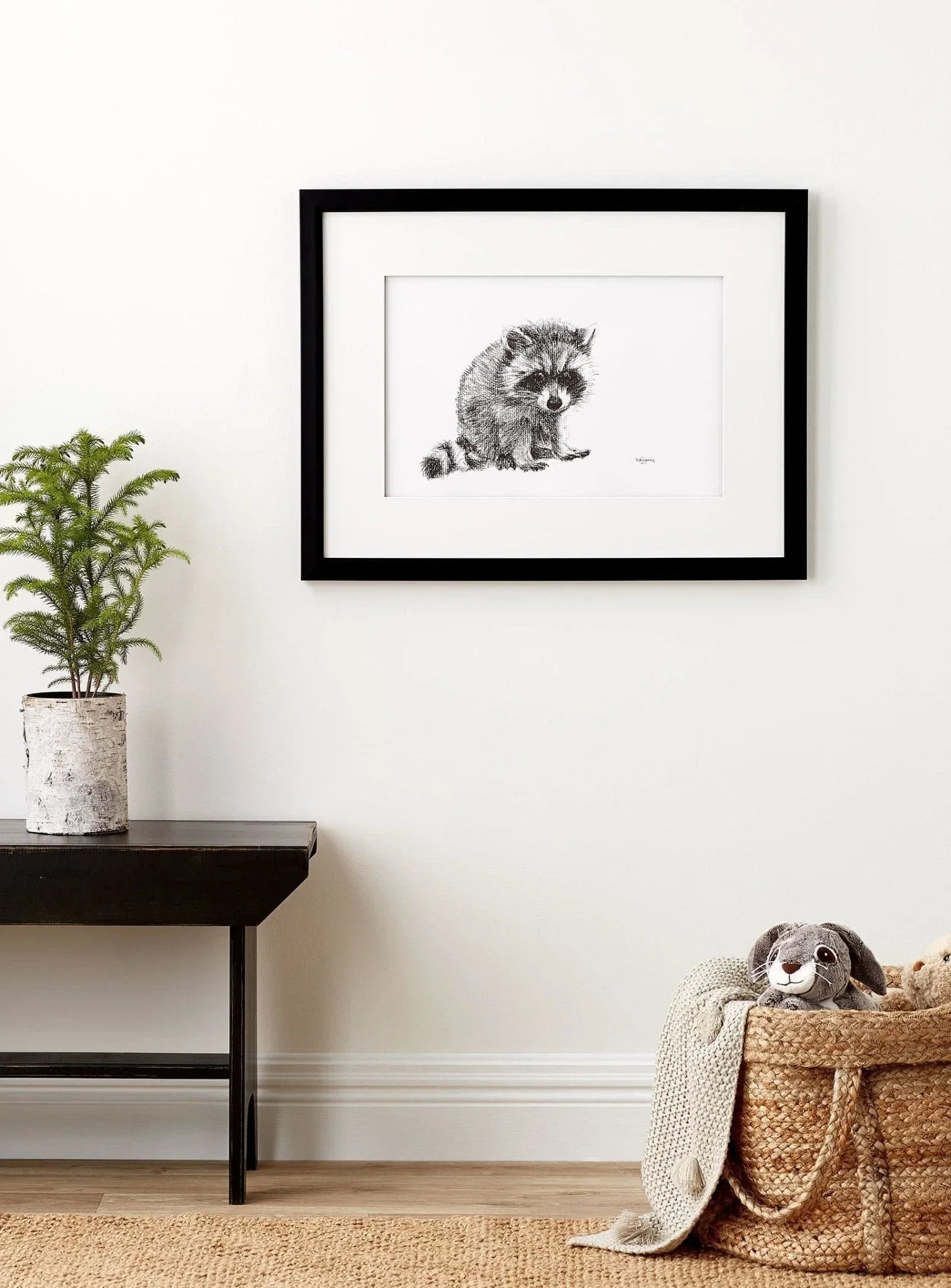 Framed baby raccoon art print on white wall above modern table and basket with plush toy