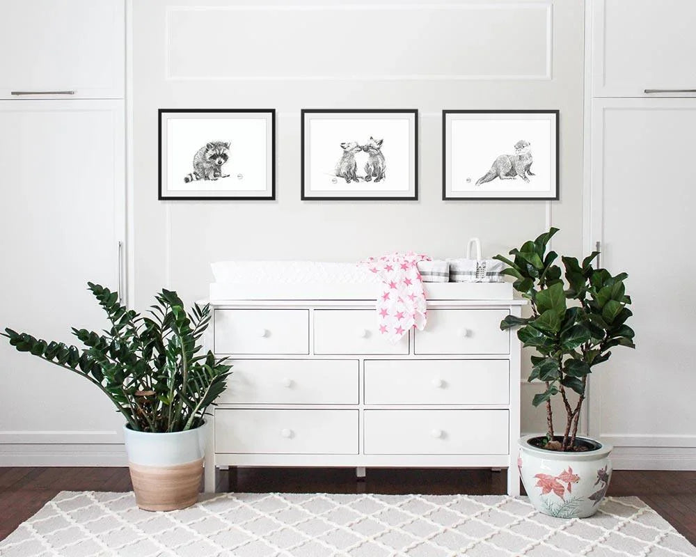 Framed baby raccoon wall art above white dresser in modern nursery with plants
