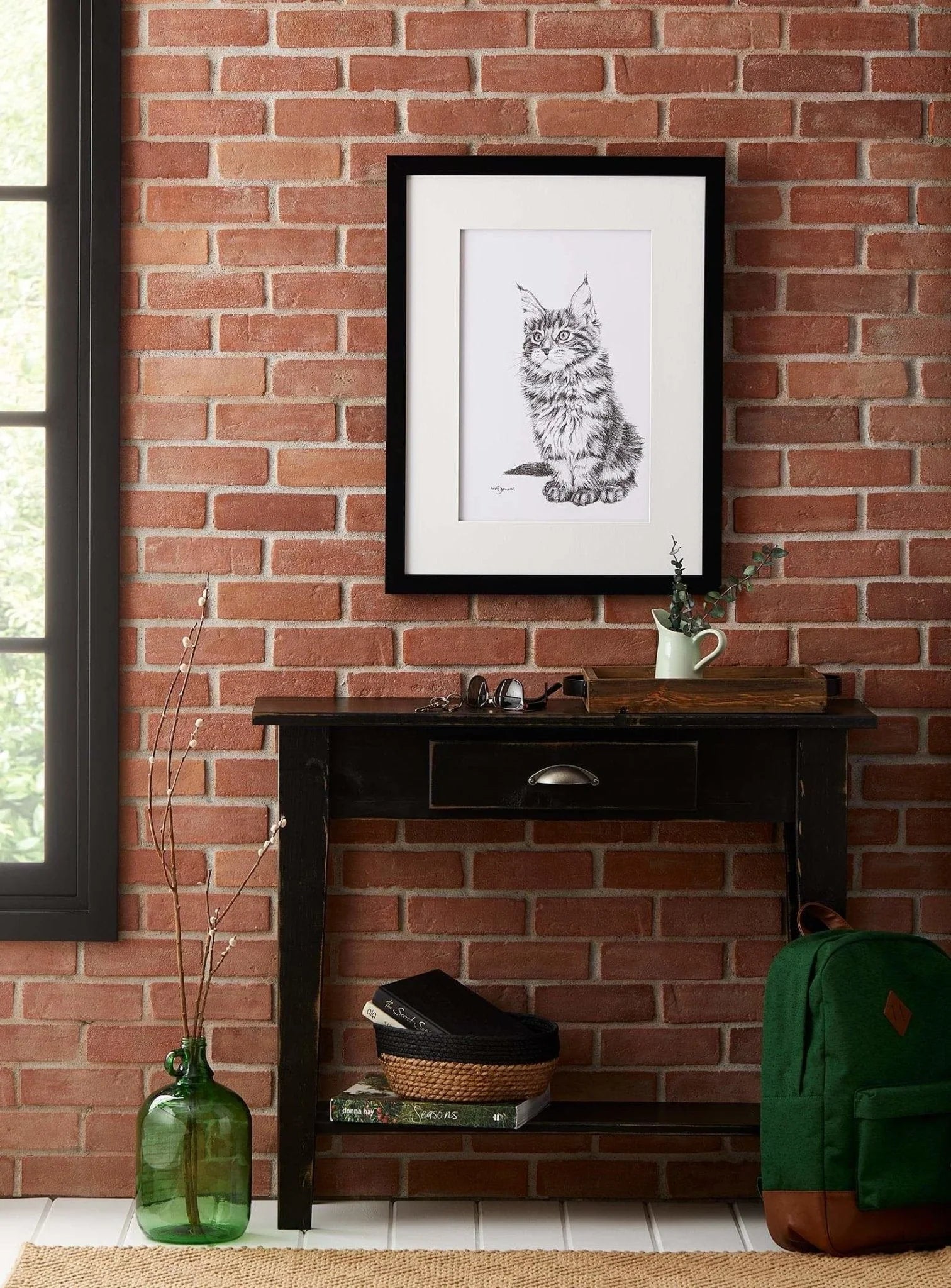 Framed cat sketch on brick wall above rustic table, green backpack, and glass vase decor