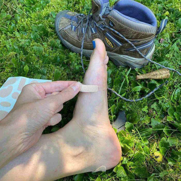 Applying pre-cut moleskin to foot outdoors near hiking boot on green grass