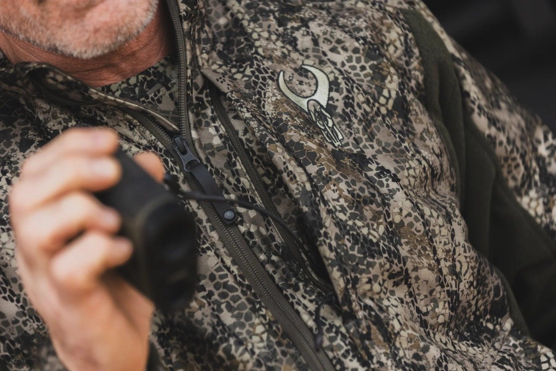Close-up of man in camouflage hunting jacket with embroidered ram skull logo holding binoculars