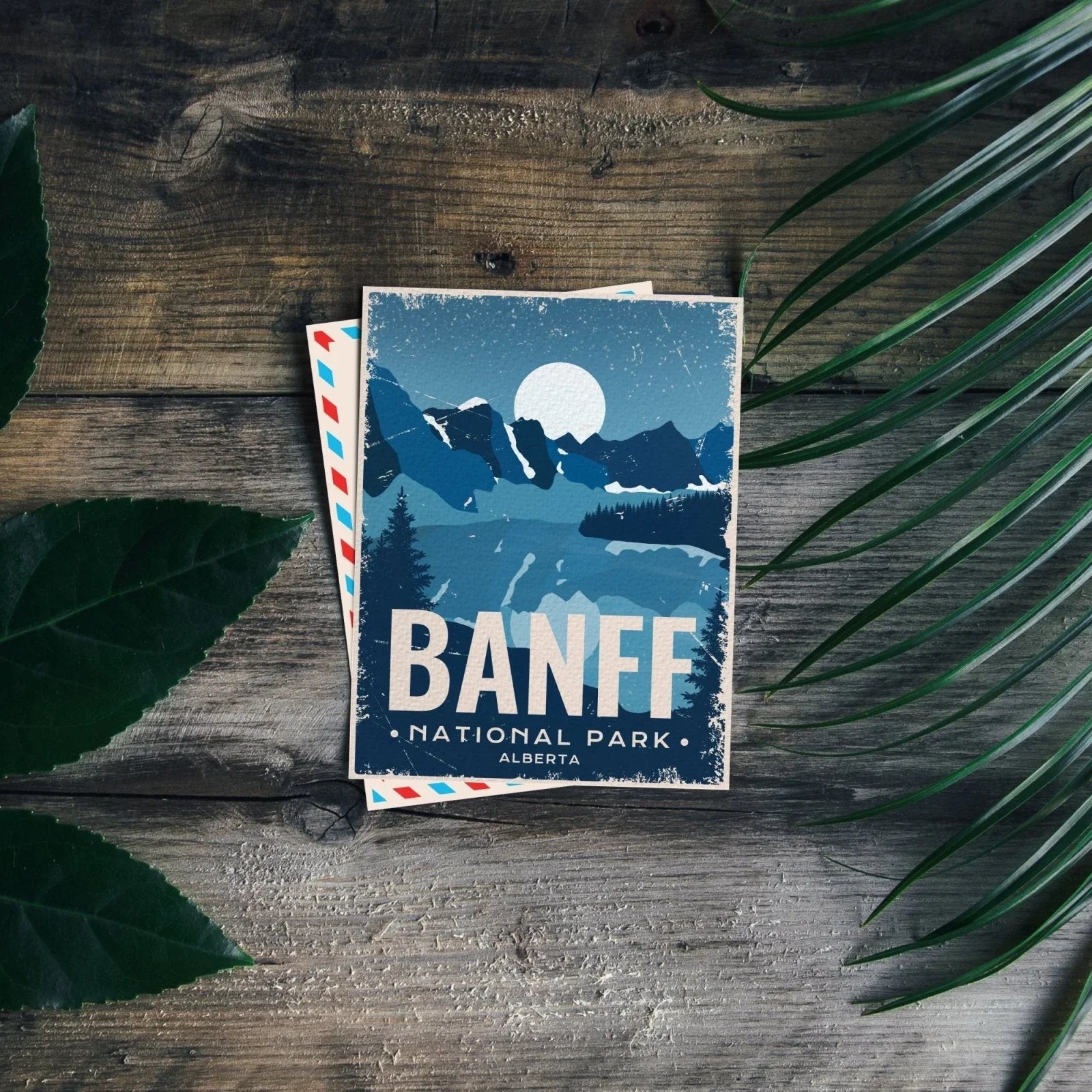 Banff National Park Alberta postcard with mountain illustration on rustic wood, leaves nearby