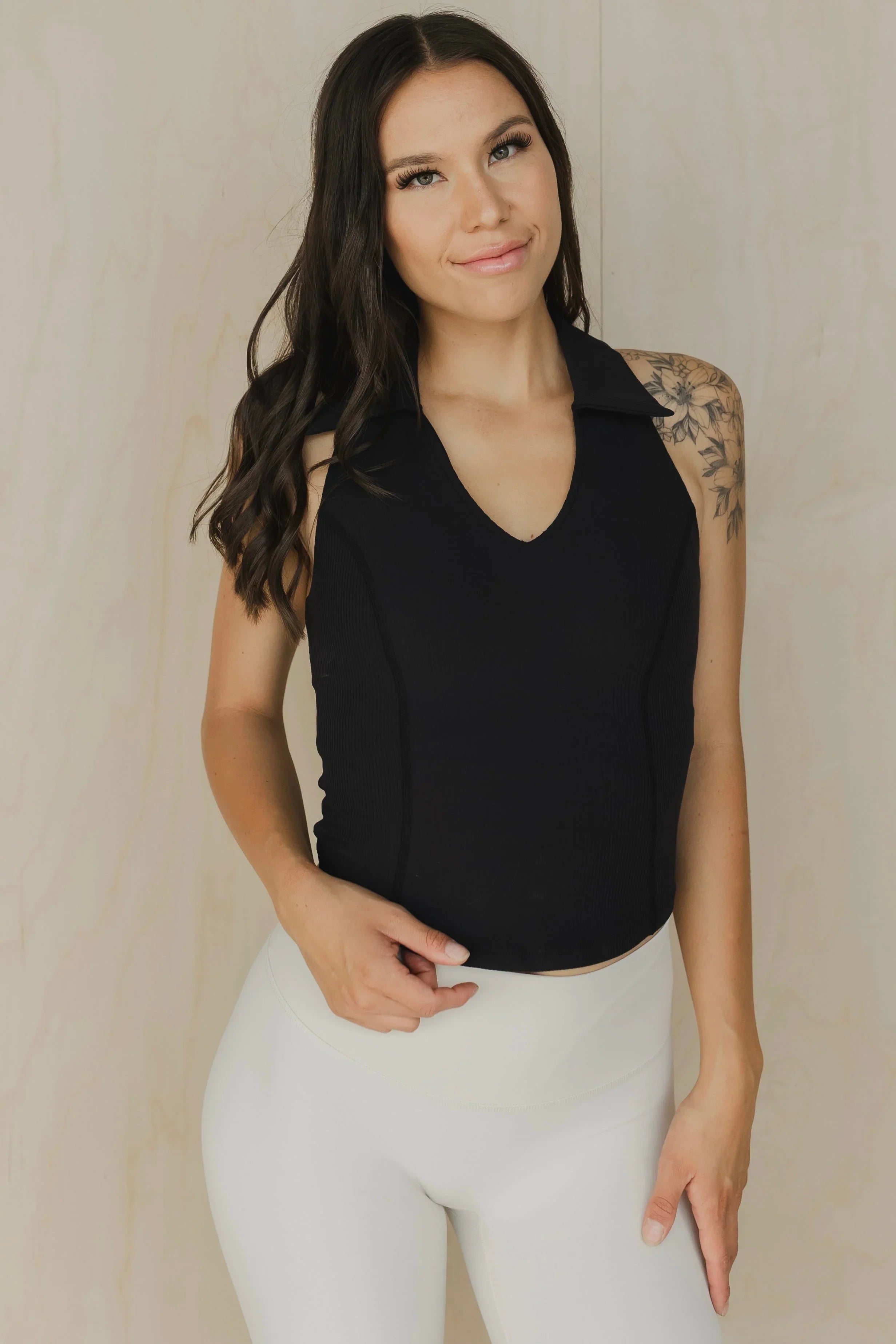 Woman in black sleeveless top and white leggings posing, floral tattoo on shoulder