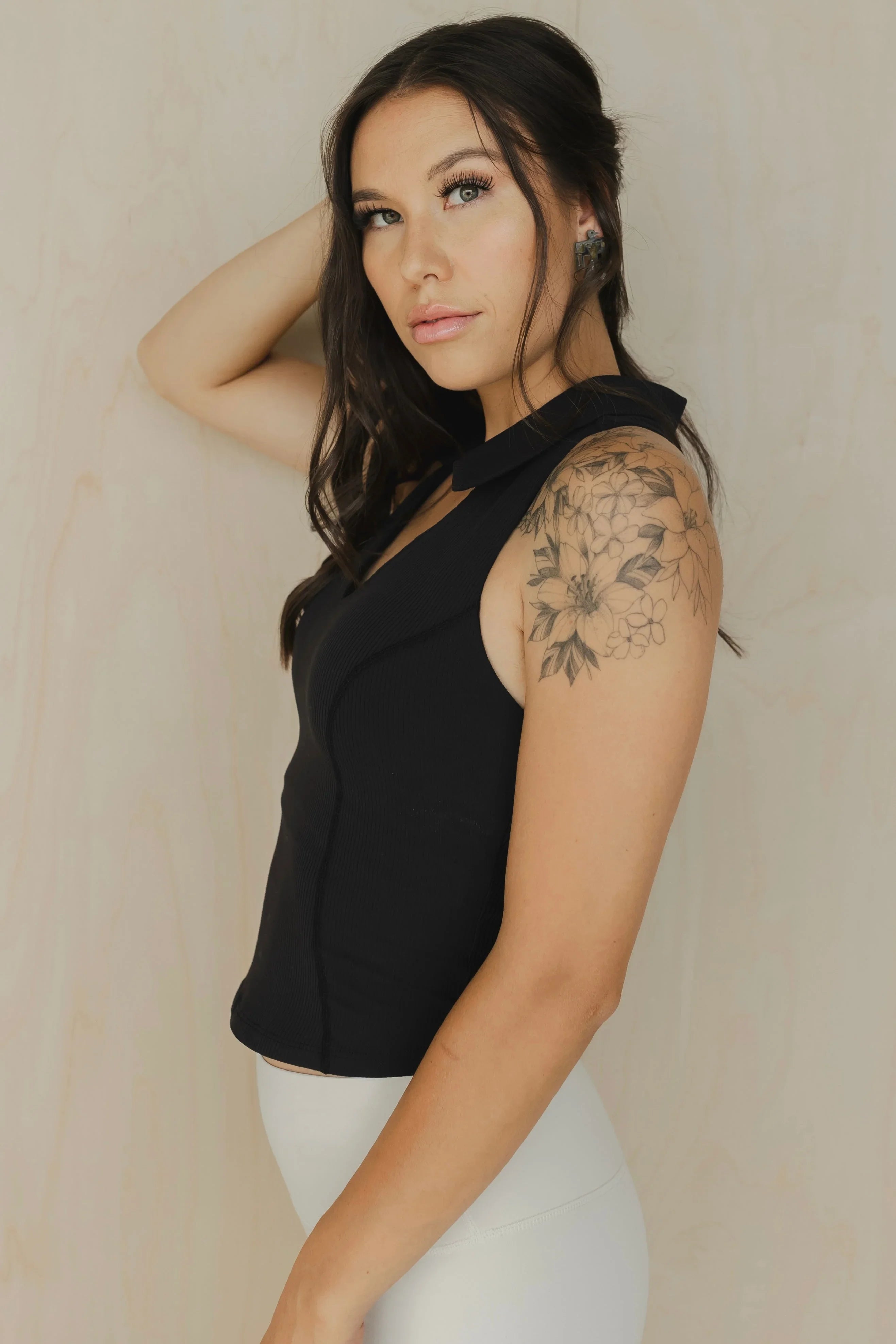 Woman in black sleeveless top, floral shoulder tattoo, posing in front of beige wall