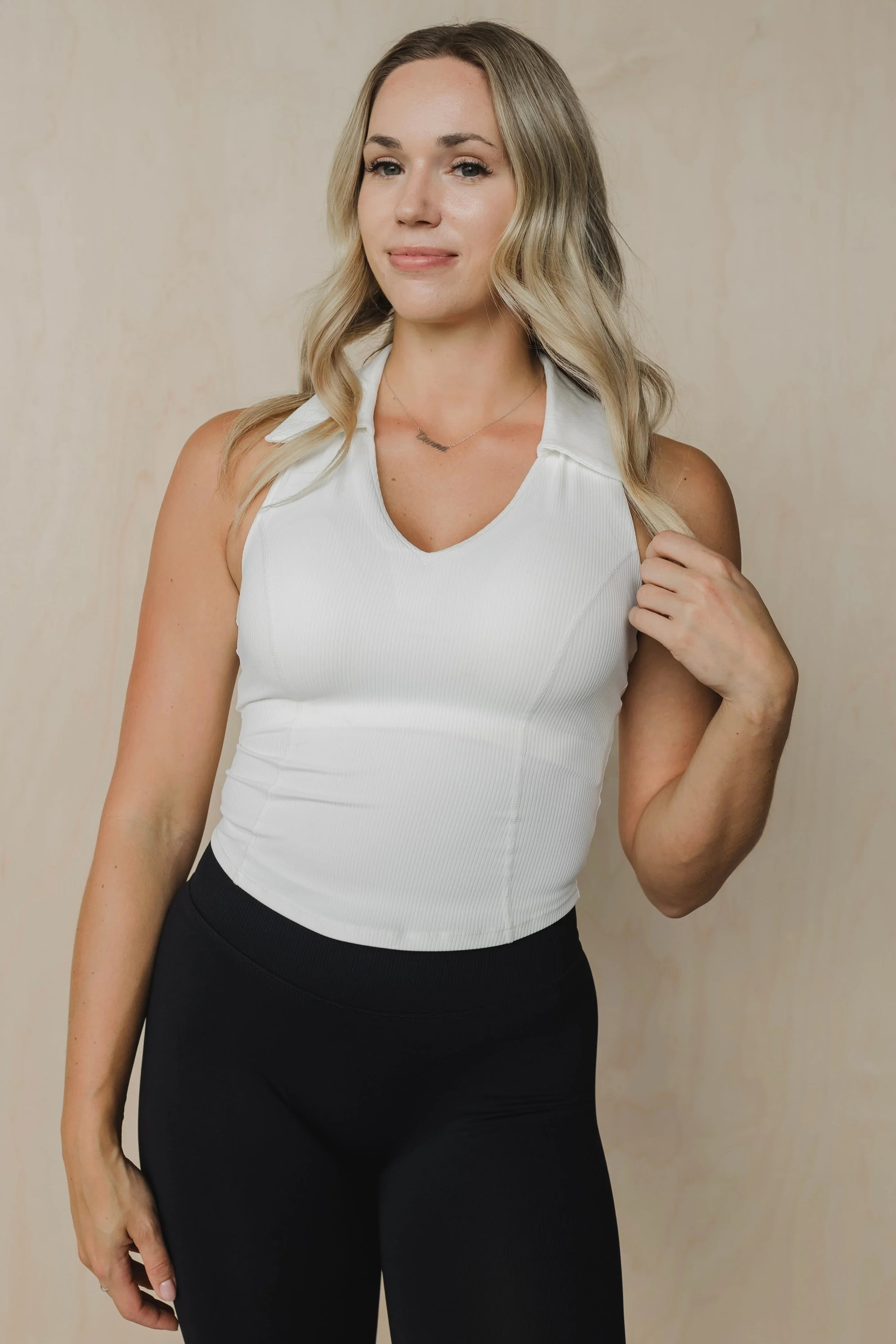 Woman in white sleeveless collared top and black leggings against a beige background