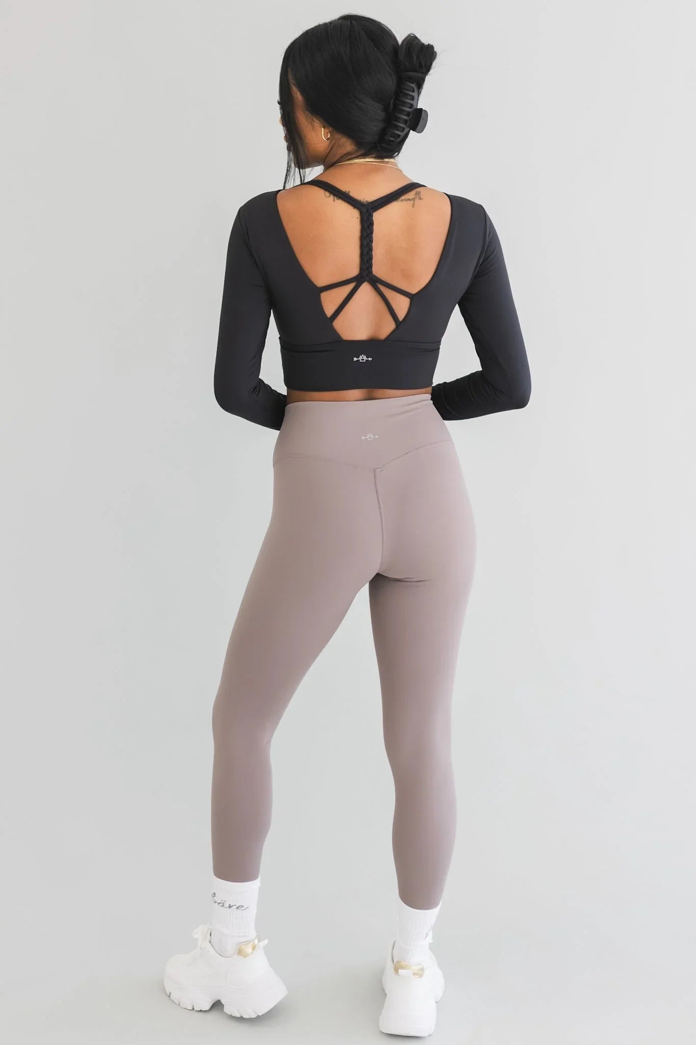 Woman in black open-back long sleeve crop top and taupe leggings with white sneakers