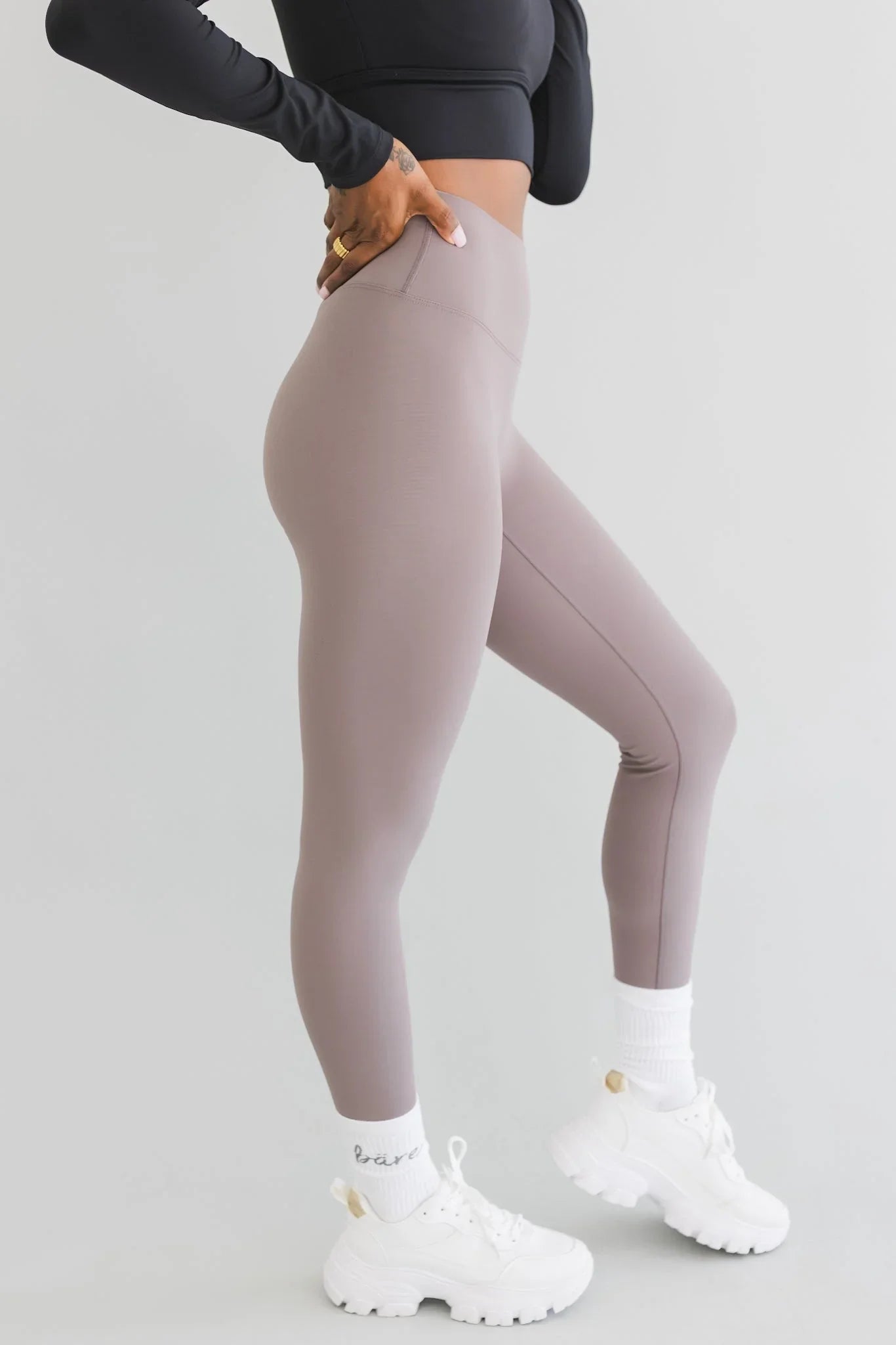 Woman in high-waisted taupe leggings, white chunky sneakers, and black crop top