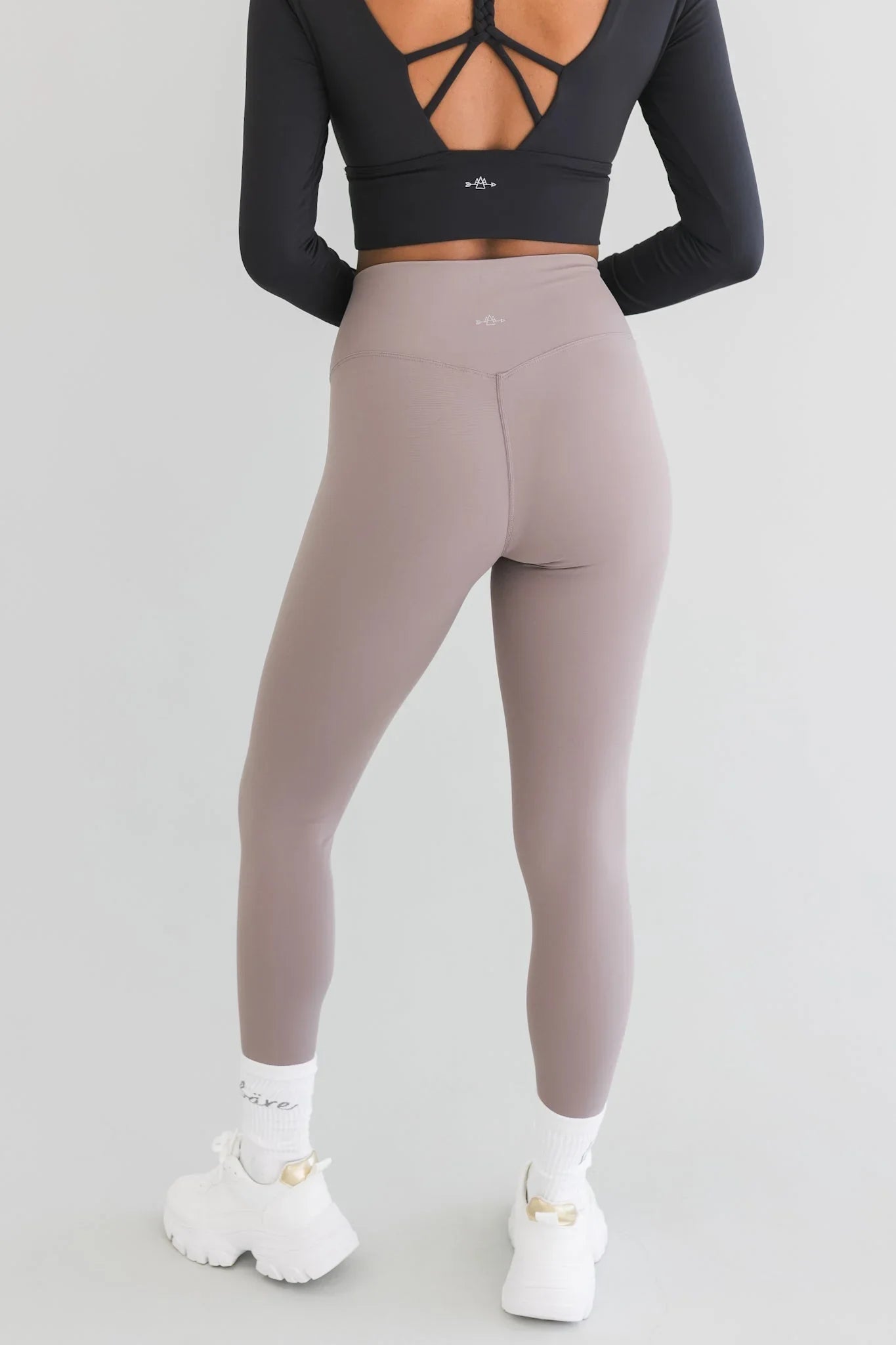 Woman in taupe high-waisted leggings and black open-back crop top, white sneakers