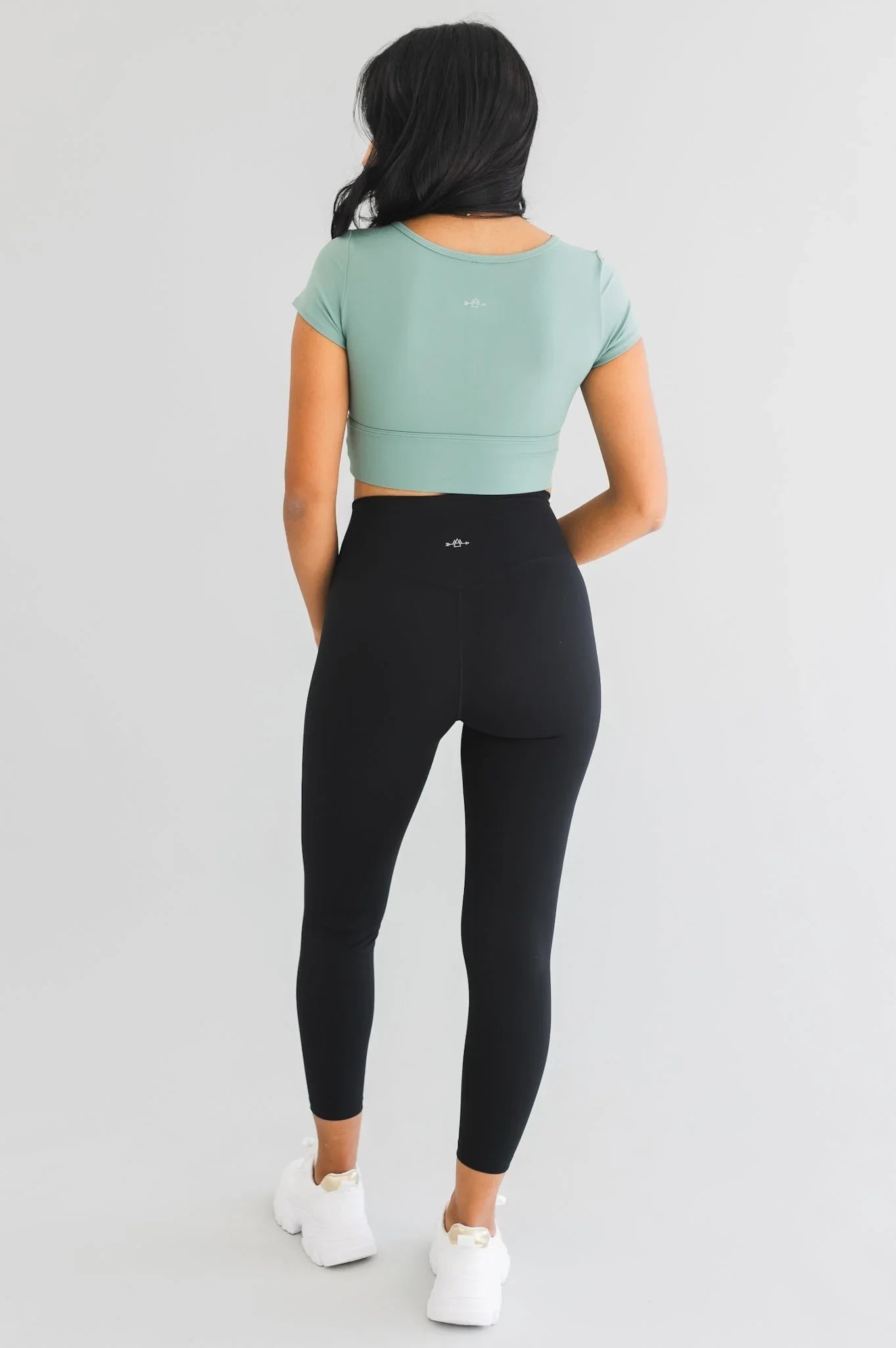 Woman in sage green crop top and black high-waisted leggings, white sneakers, studio background