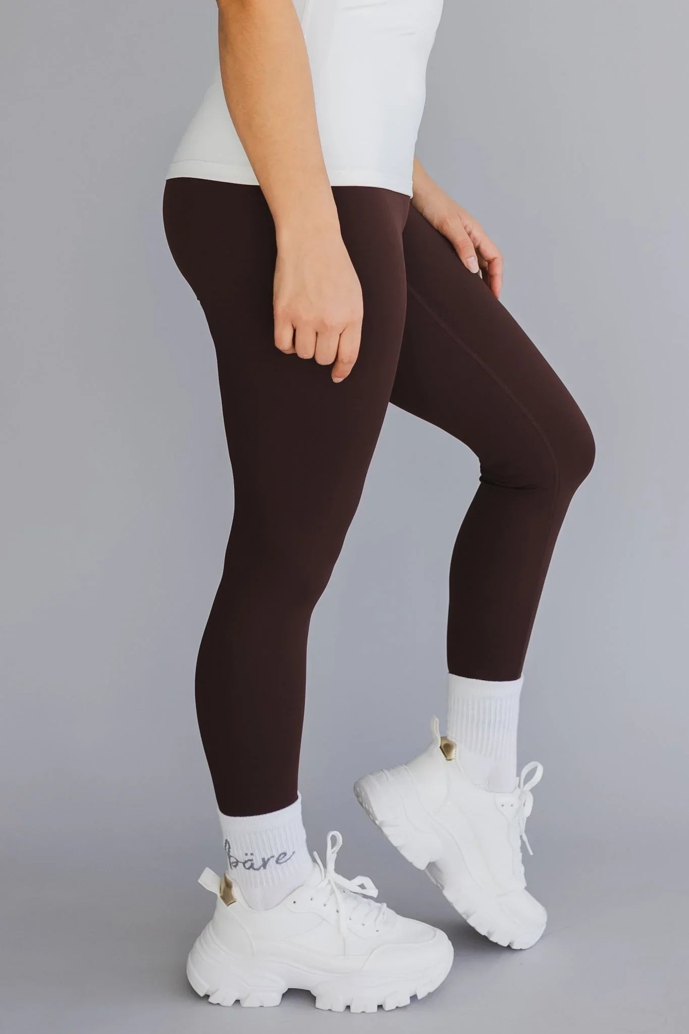 Woman wearing brown leggings, white top, and chunky white sneakers against gray background