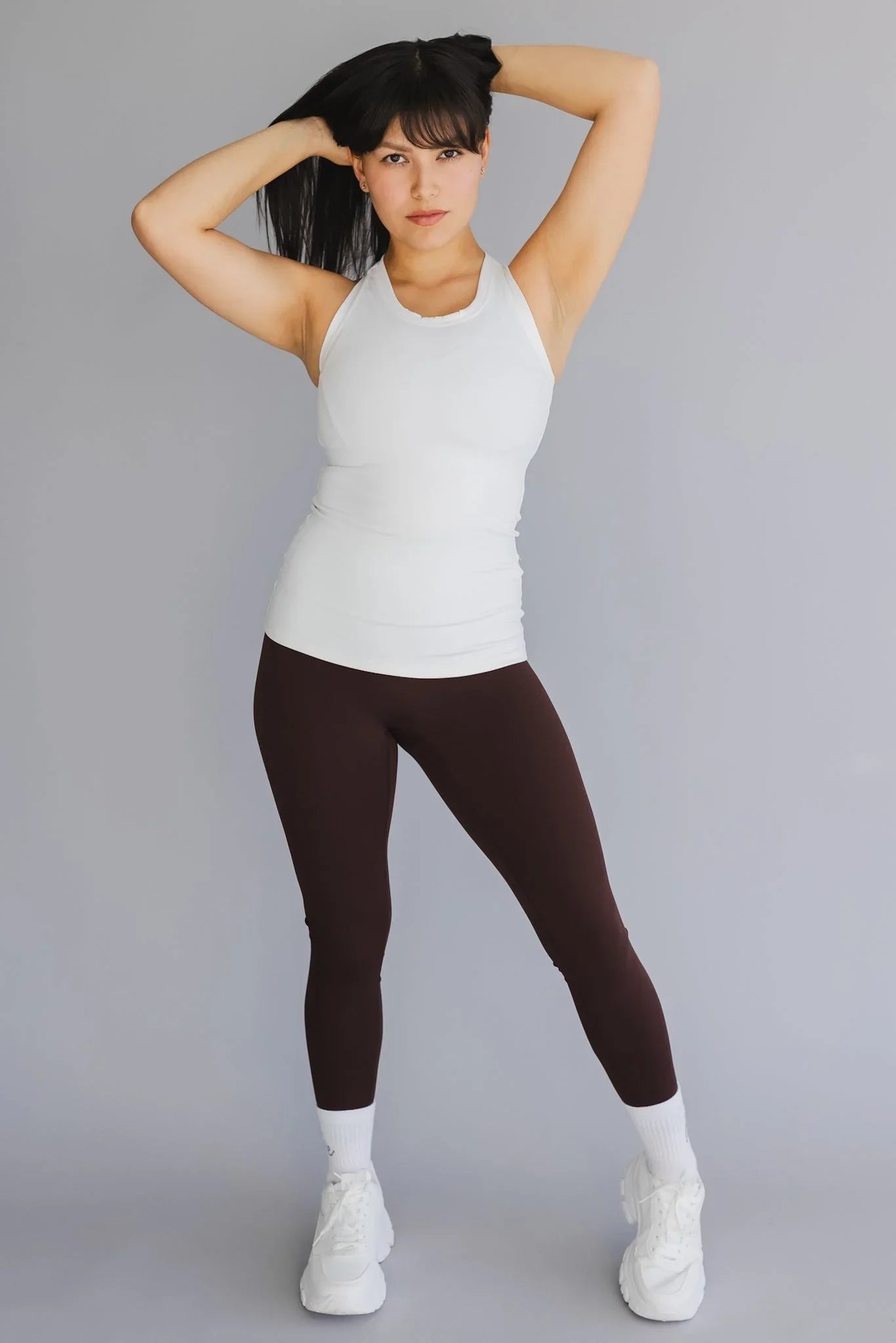 Woman in white tank top and brown leggings posing against gray background, athletic wear