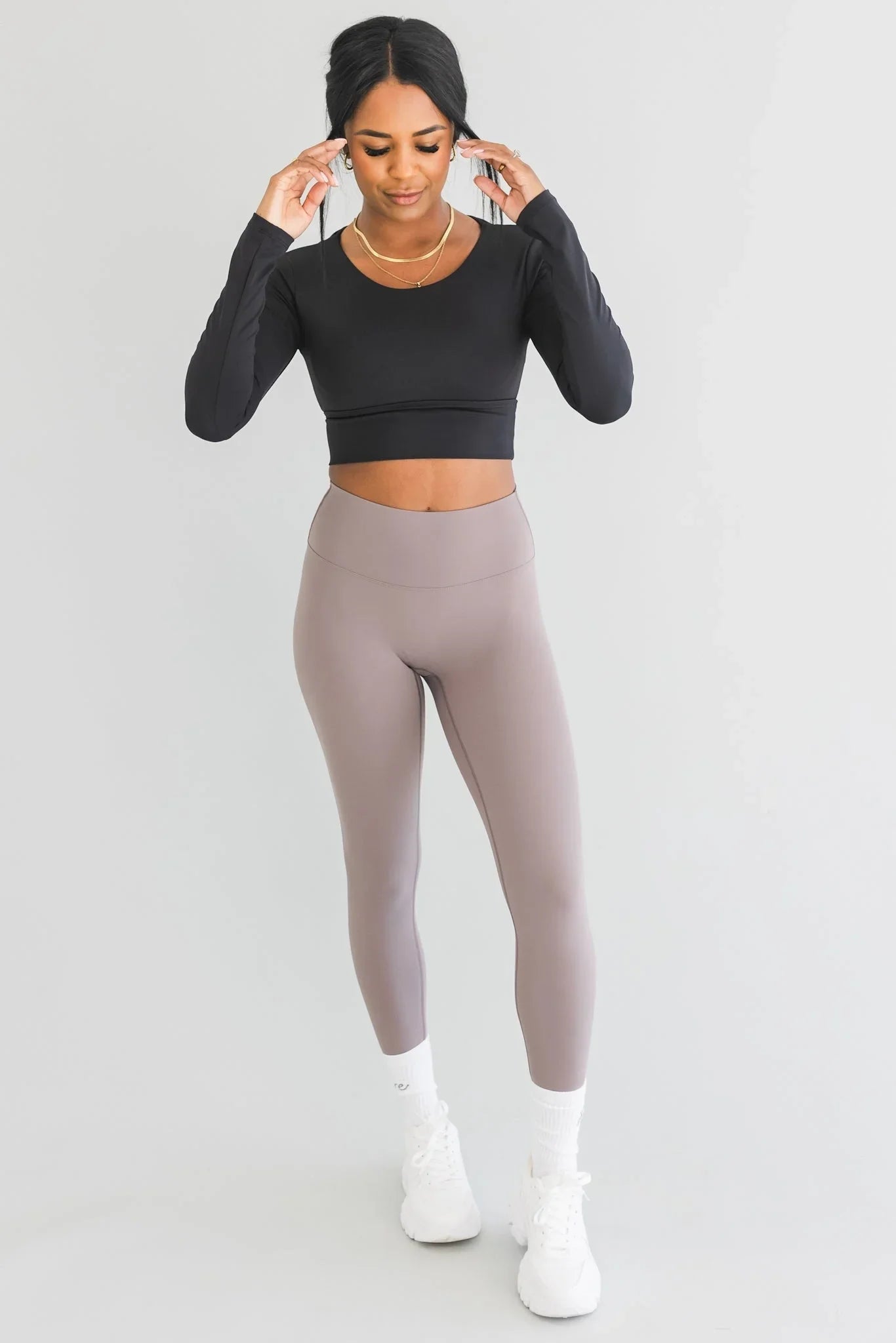Woman in black long sleeve crop top and taupe leggings, white sneakers, posing indoors