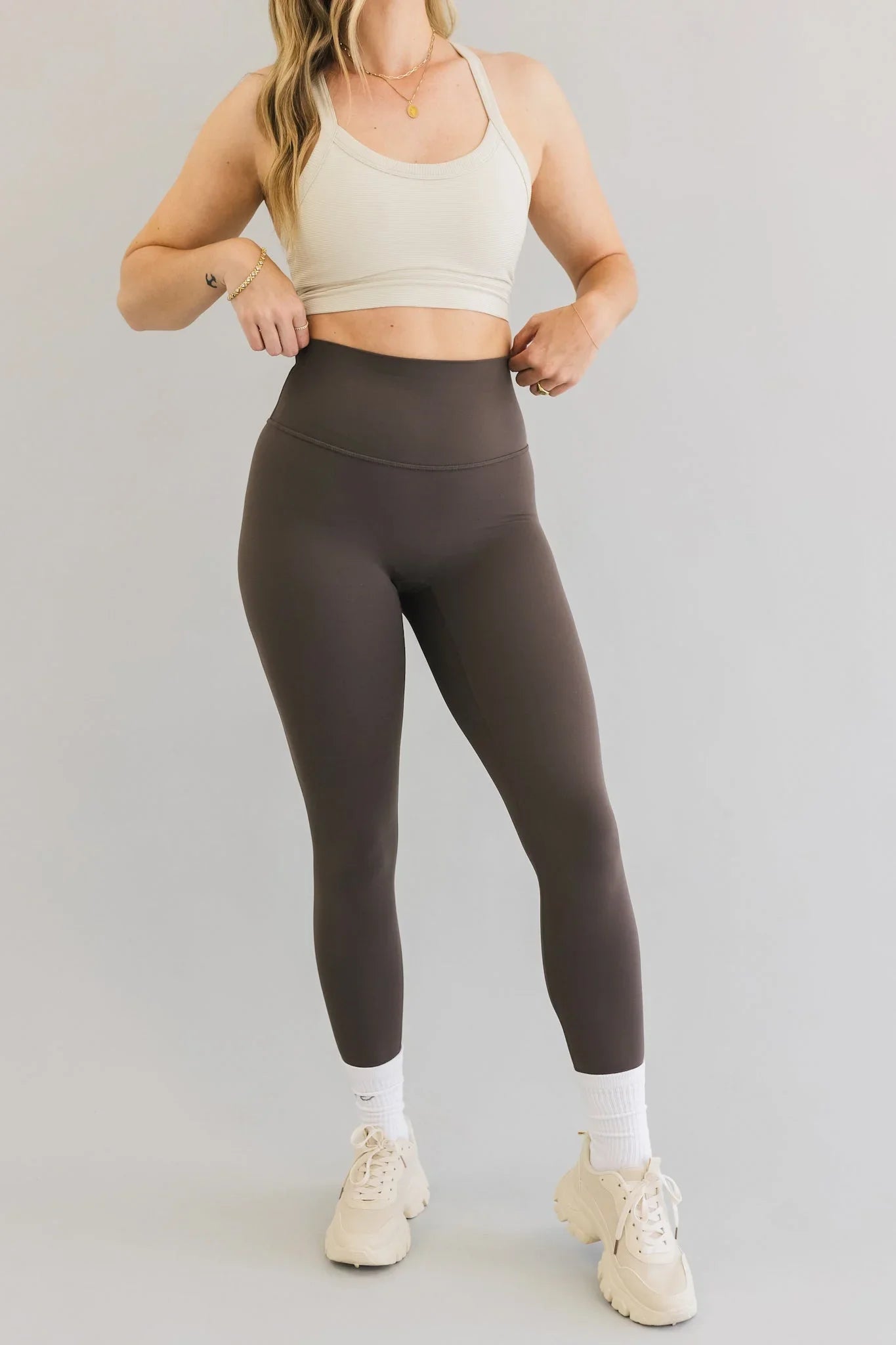 Woman modeling high-waisted brown leggings, cream sports bra, and chunky sneakers
