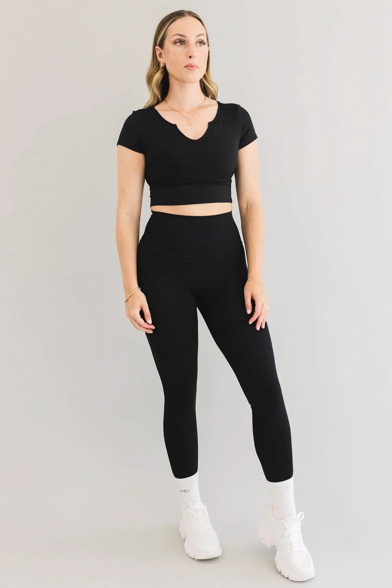 Woman wearing black crop top and high-waisted leggings, white sneakers, standing on gray background