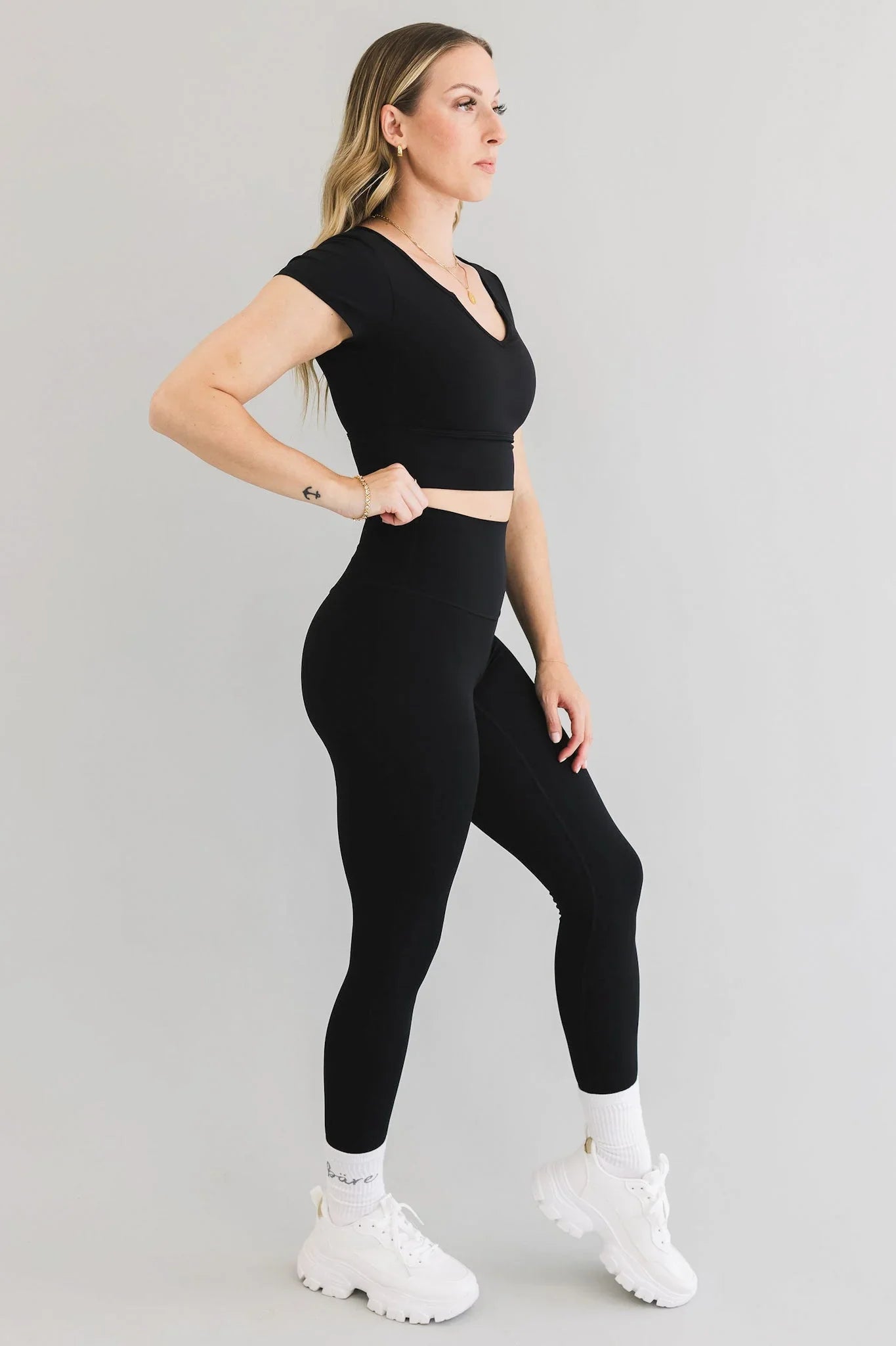 Woman modeling black activewear set with white chunky sneakers against plain background