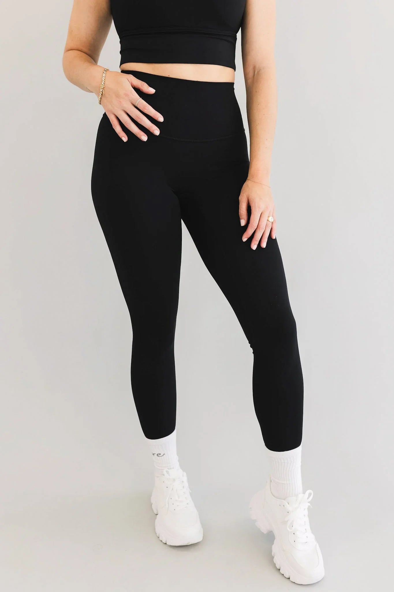 Woman wearing black high-waisted leggings, black crop top, white sneakers, and gold jewelry