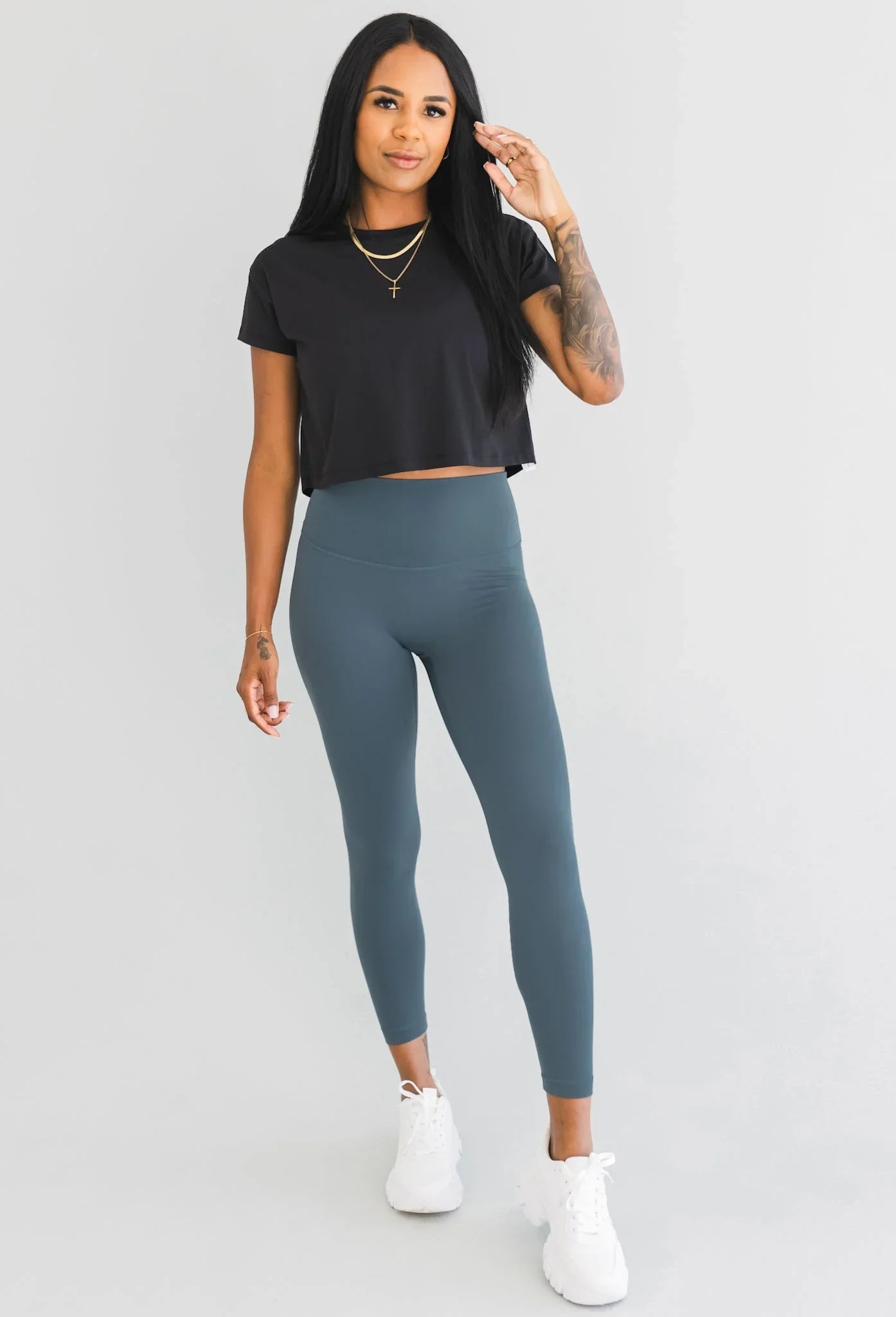 Woman in black crop top, blue high-waisted leggings, and white sneakers on white background