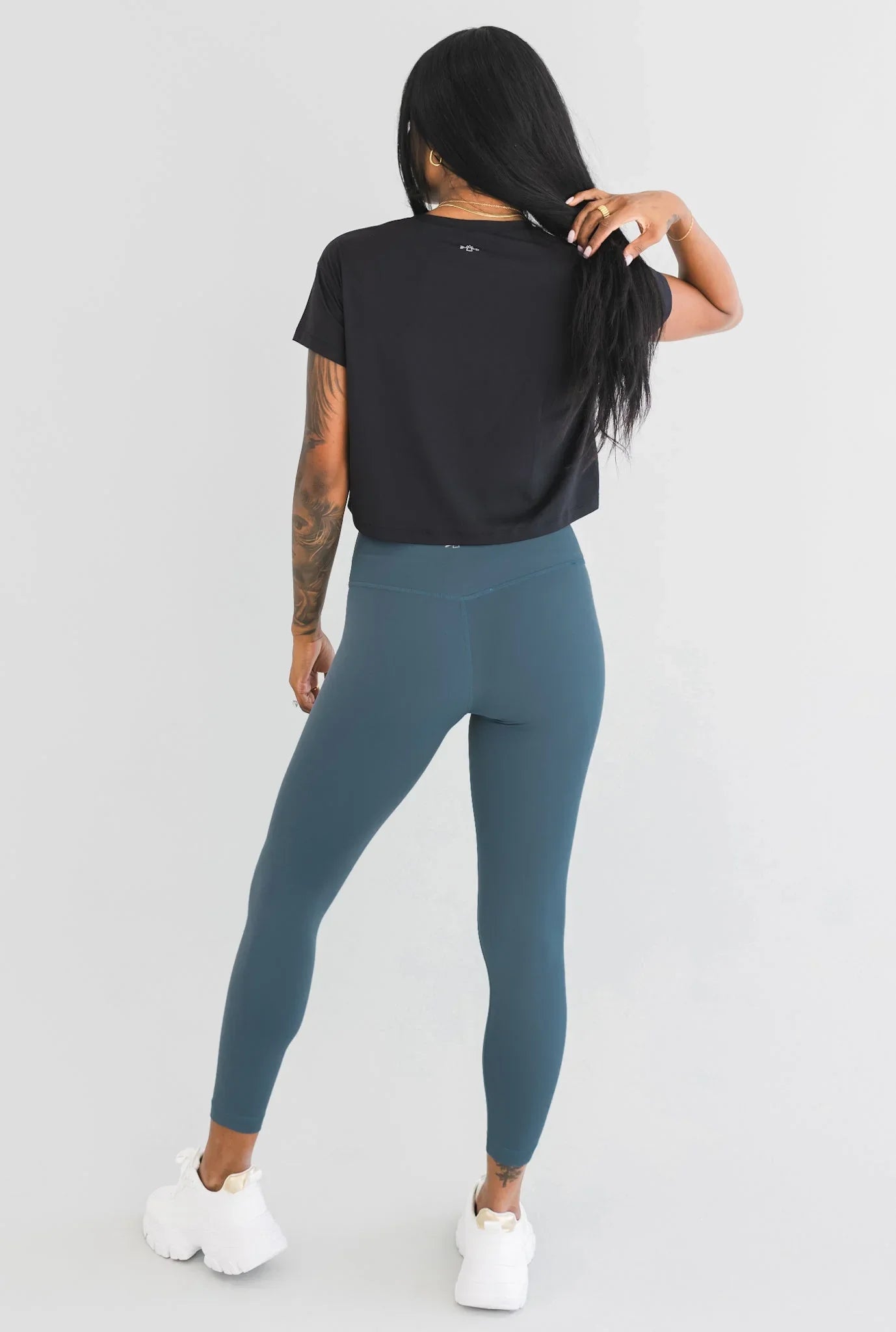 Woman in black t-shirt, blue leggings, and white sneakers posing against light background