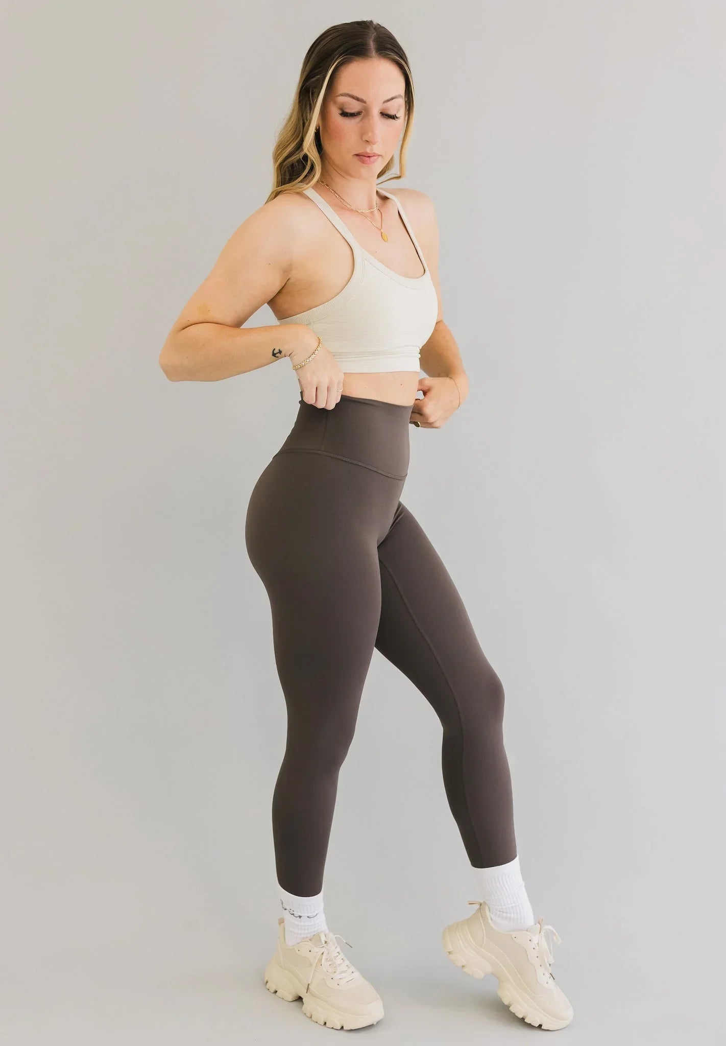 Woman in cream sports bra and high-waisted brown leggings posing on grey background