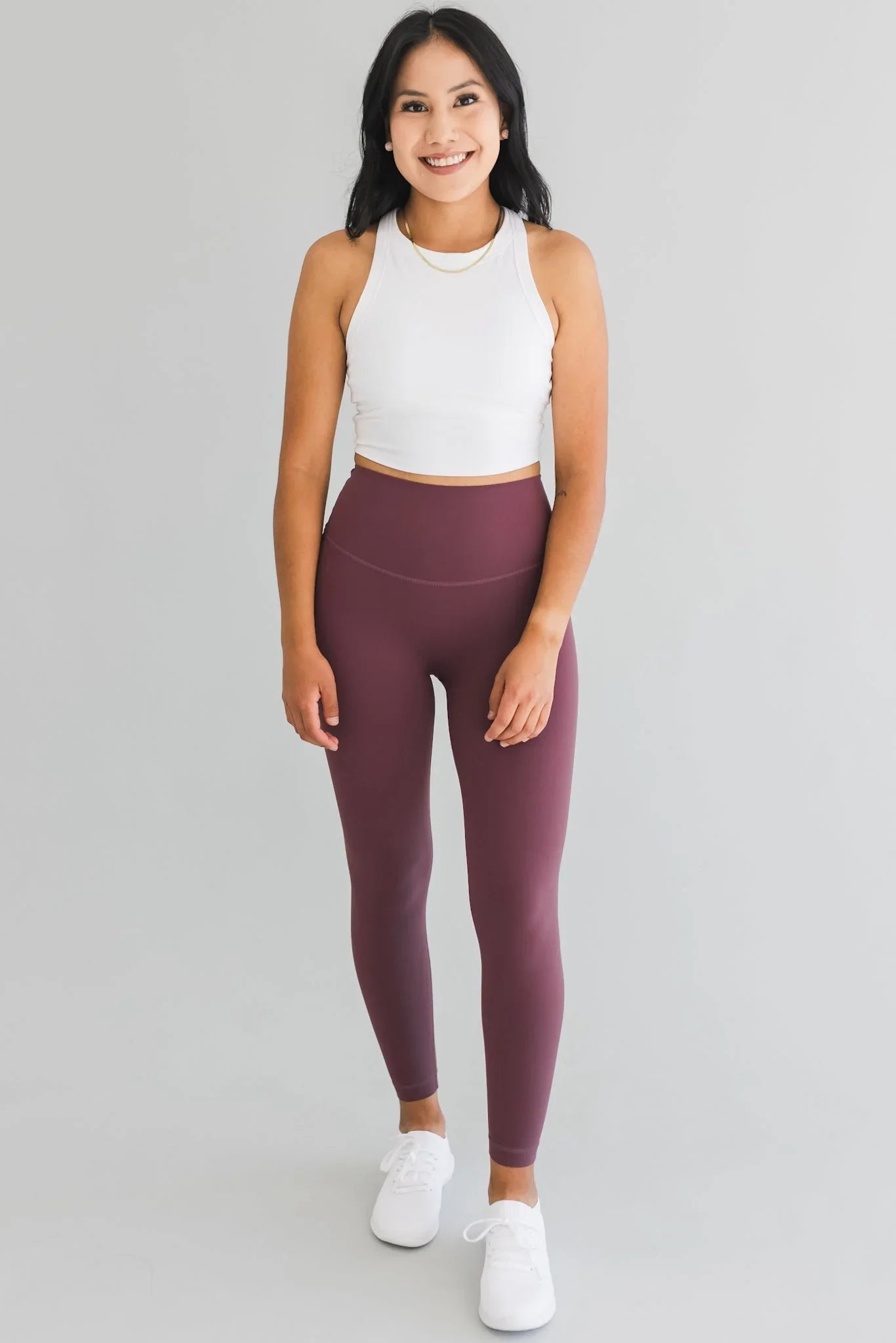 Woman in white crop top and high-waisted mauve leggings, standing against gray background