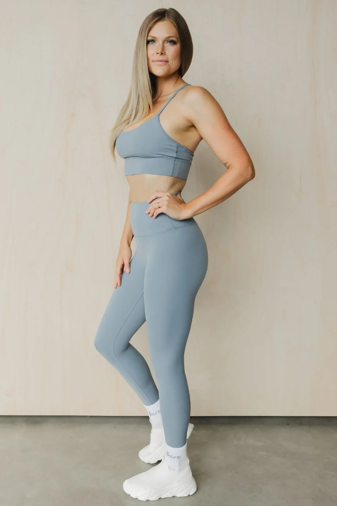 Woman in gray sports bra and leggings with white sneakers posing against beige wall